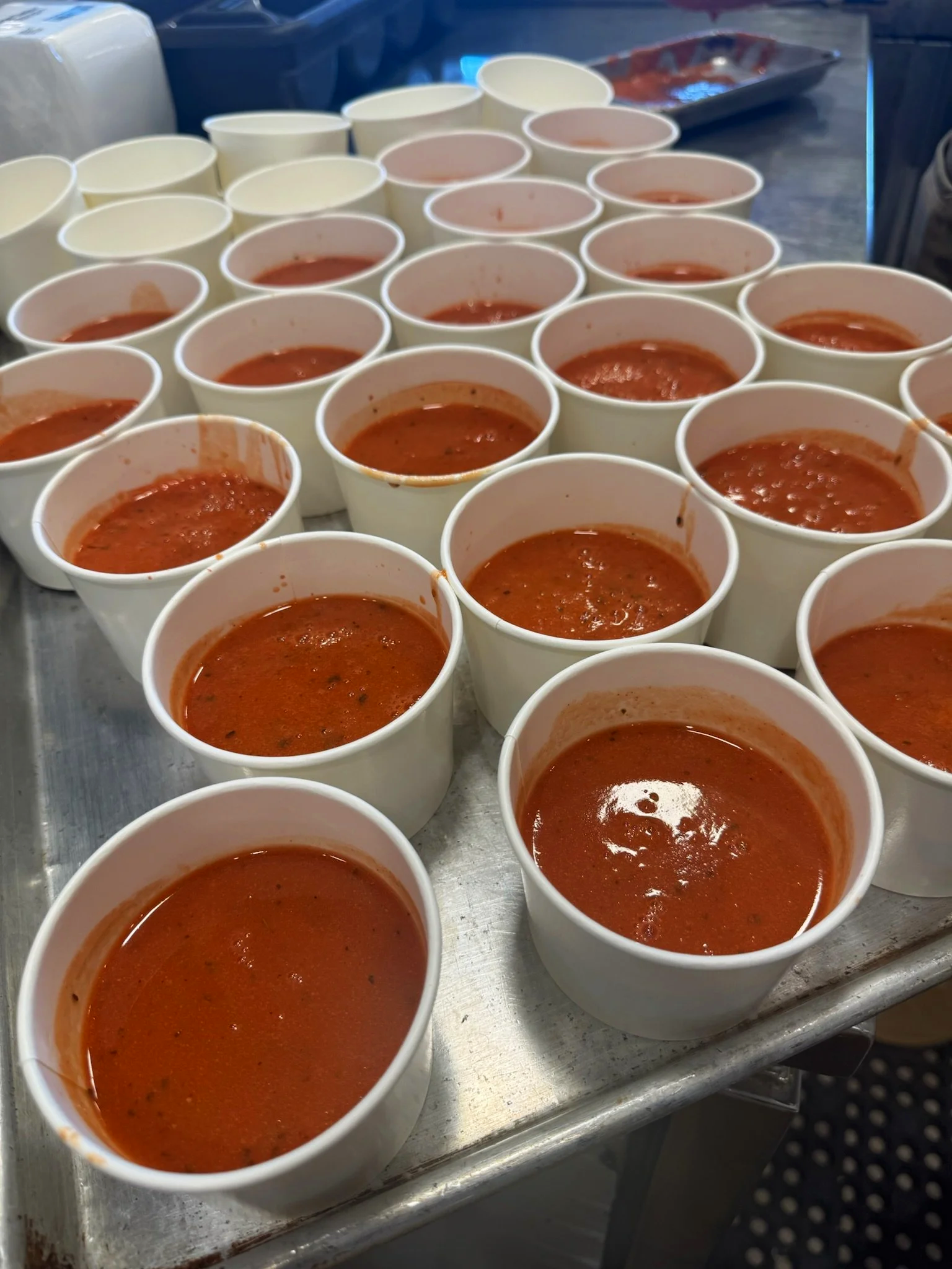 Multiple cups of tomato soup on a metal counter, with some cups empty or nearly empty.