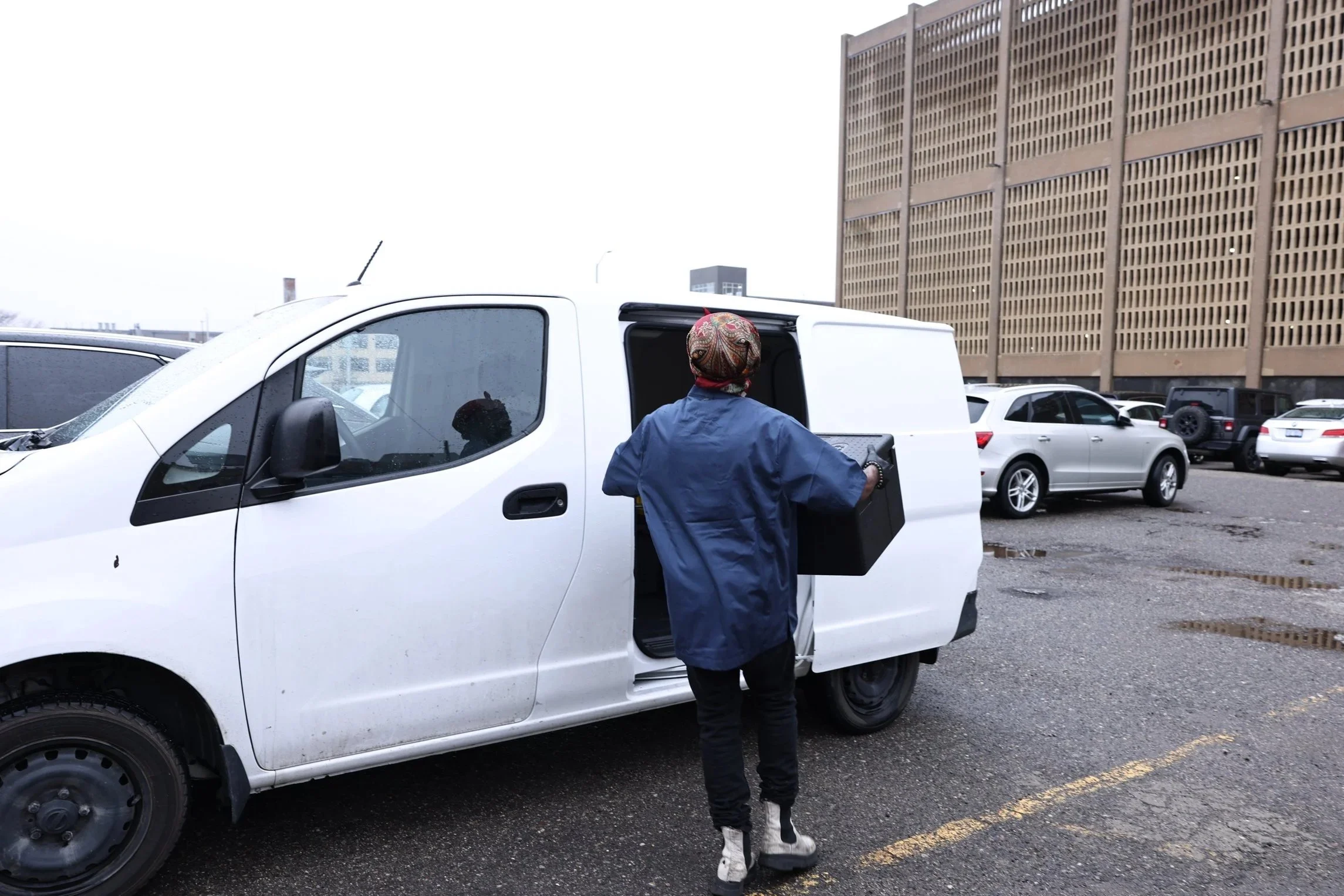 A person wearing a colorful headscarf and blue jacket loading a black box into a white cargo van in a parking lot, with several other parked cars and a large brown building in the background.
