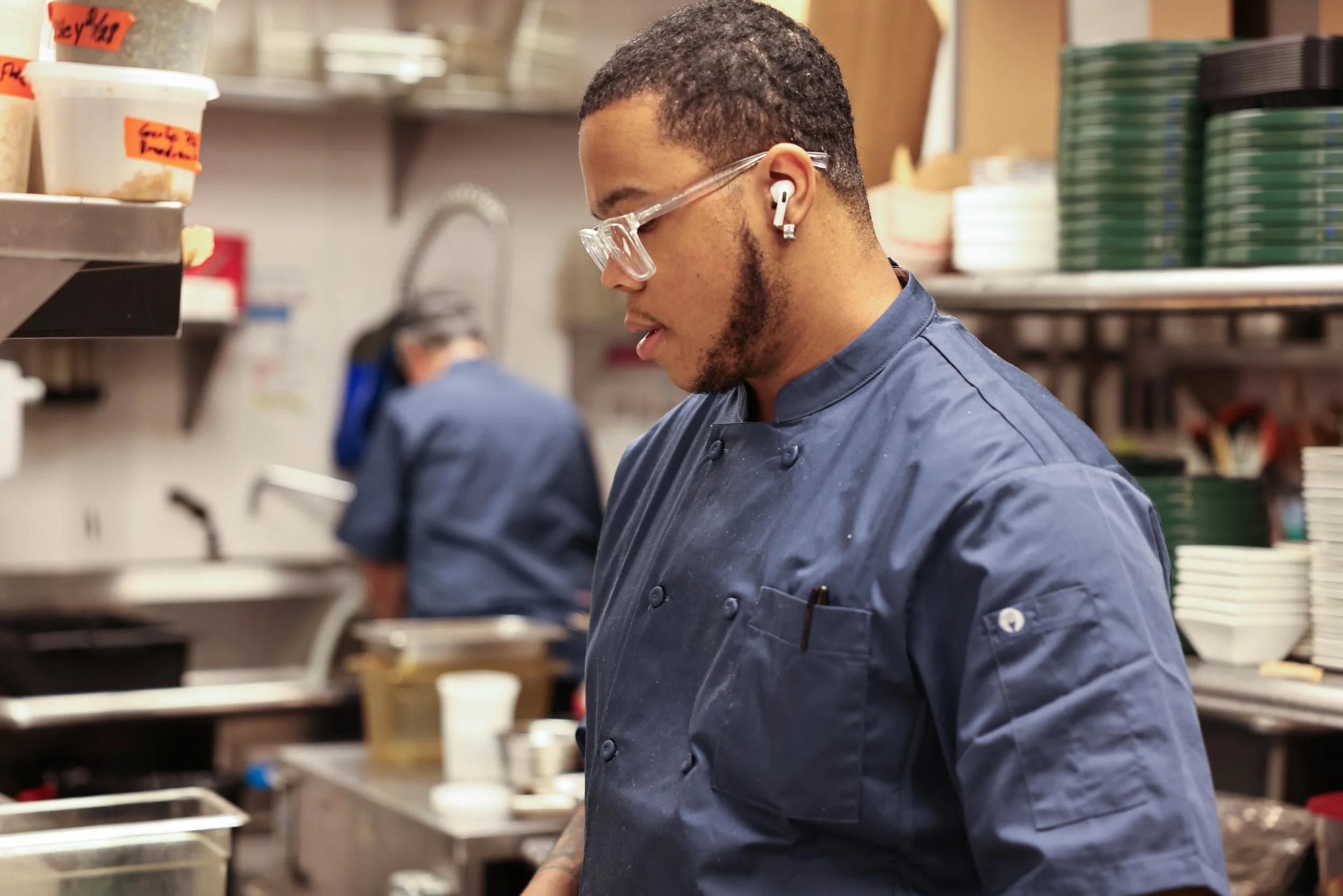 A male chef in a navy blue uniform is working in a commercial kitchen, wearing glasses and wireless earbuds, with another chef blurred in the background.