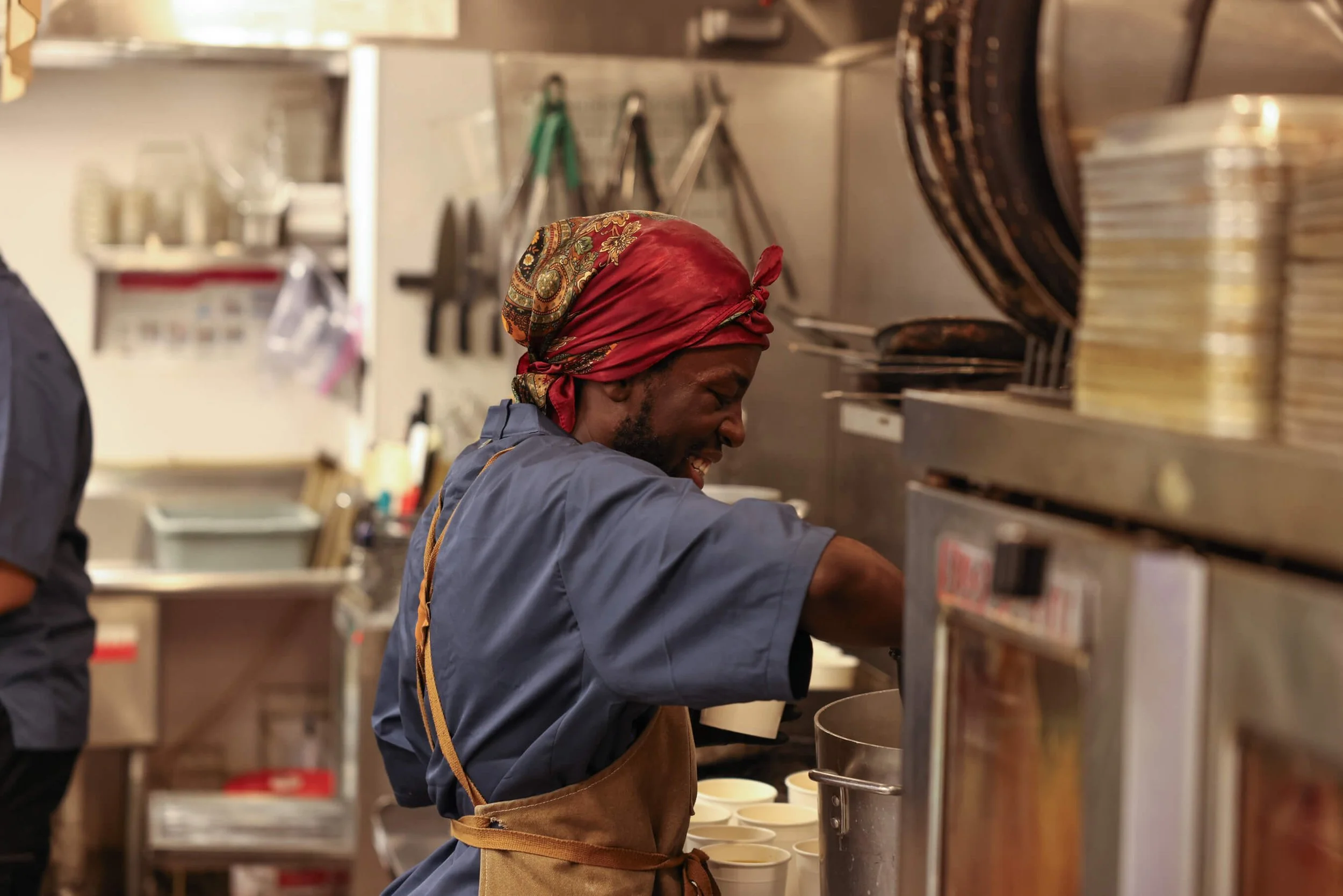 A male chef in a kitchen wearing a colorful headscarf, a blue chef's jacket, and a brown apron, preparing food.