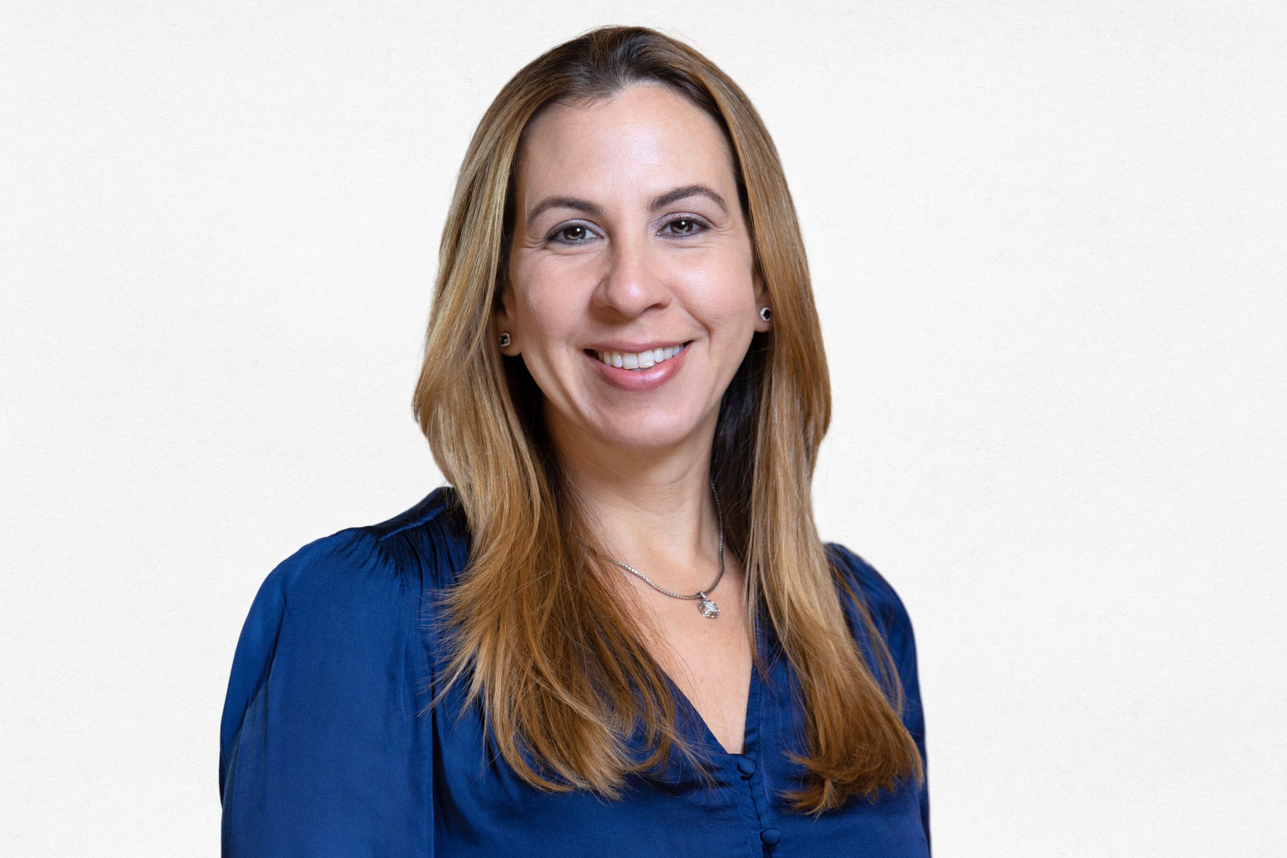 A woman with long, light brown hair smiling, wearing a blue blouse and a necklace, against a plain light background.