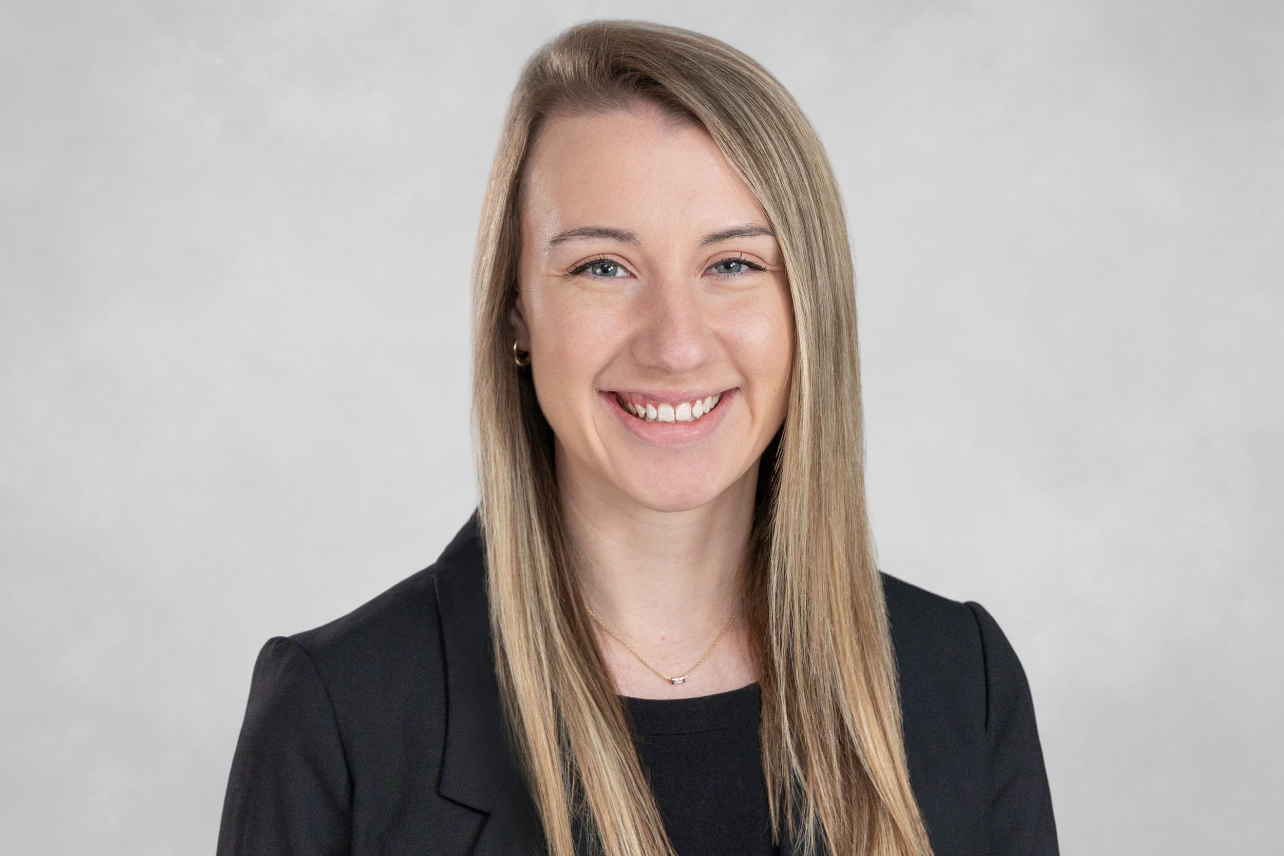 A professional woman with long blonde hair, wearing a black blazer and a delicate necklace, smiling against a light grey background.