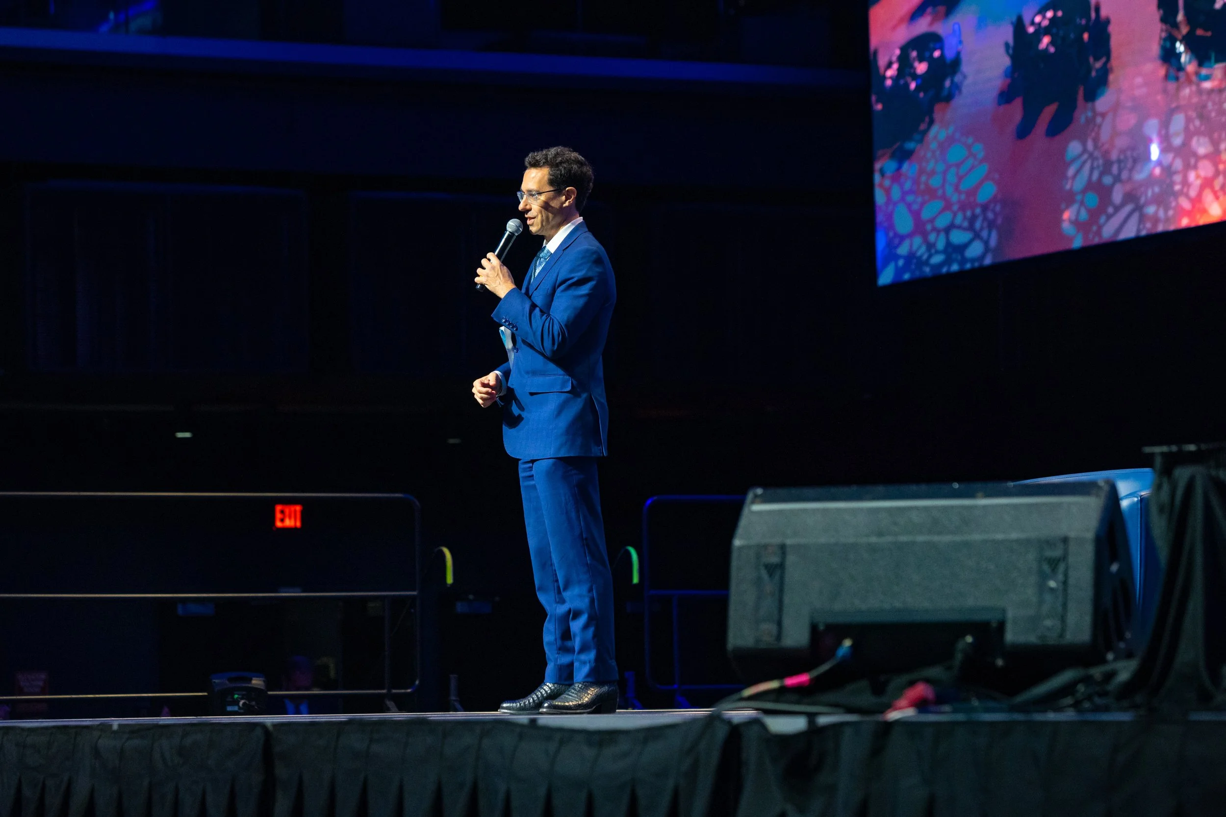 A man in a dark suit and glasses giving a presentation at a conference, standing near a podium with a logo featuring a red heart inside a geometric shape, and an American flag in the background.