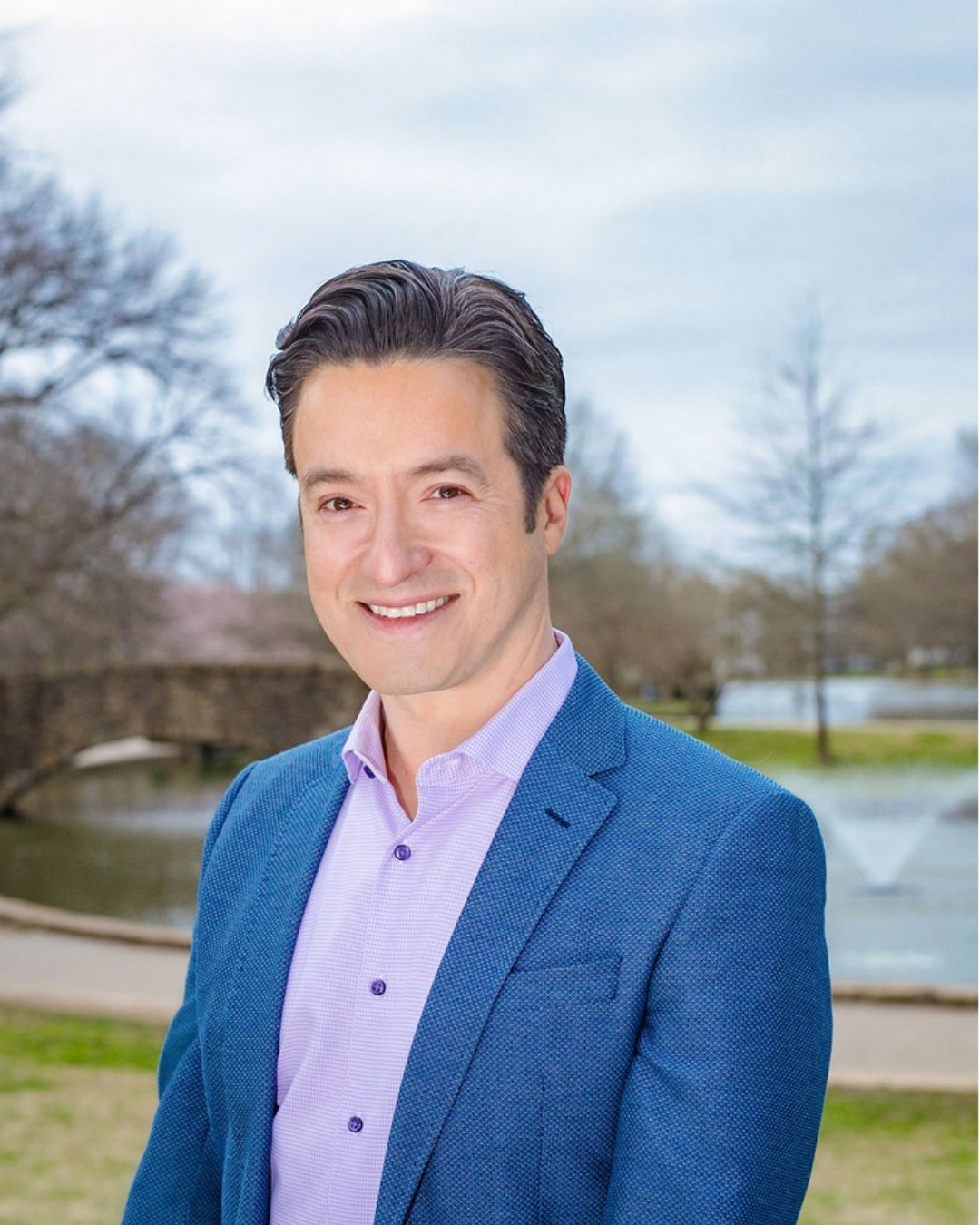 A smiling man in a blue suit jacket and light purple shirt standing outdoors near a pond with trees and a stone bridge in the background.