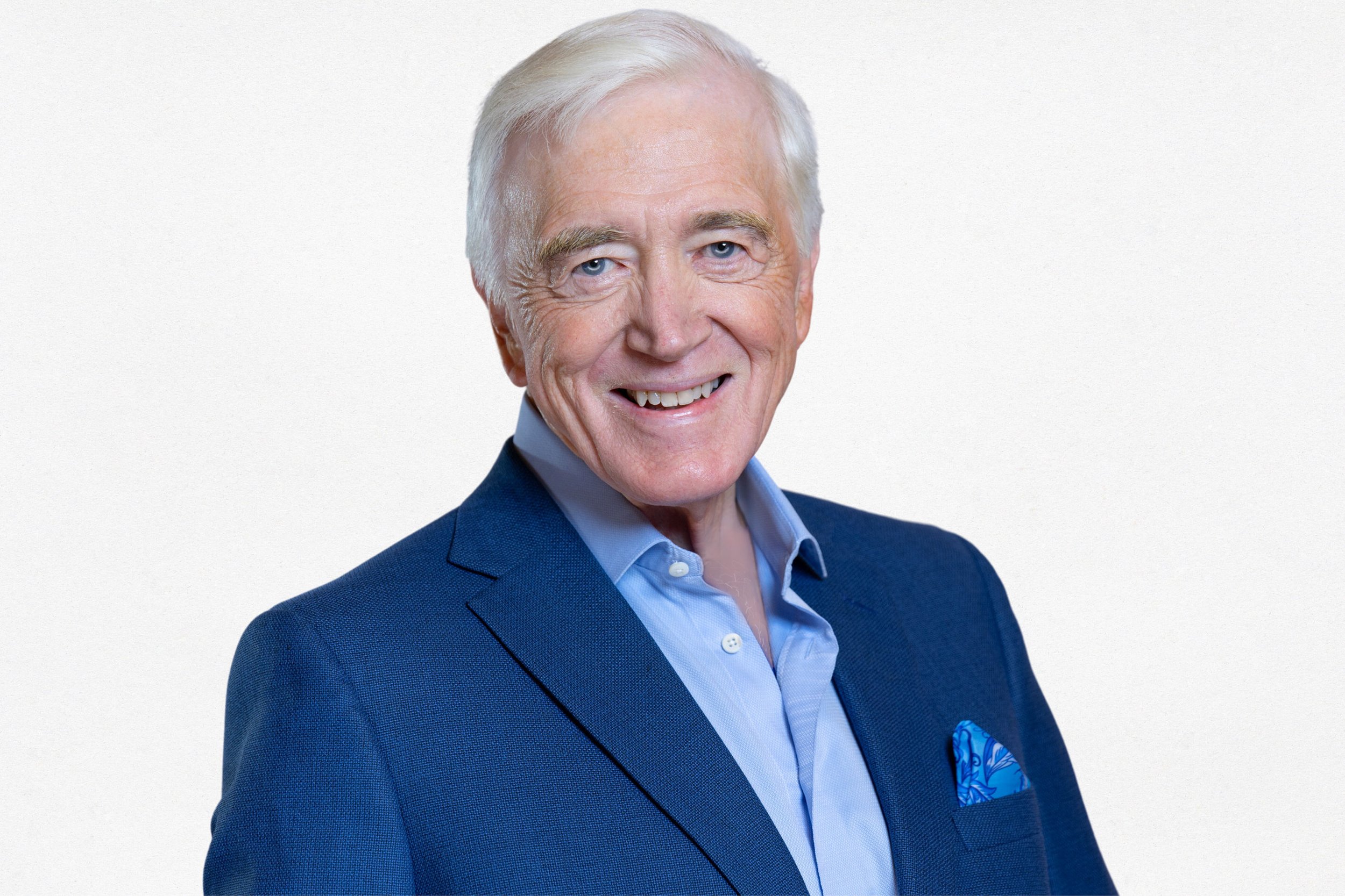 A smiling elderly man with white hair, wearing a blue suit jacket, a light blue dress shirt, and a matching blue pocket square, against a plain white background.