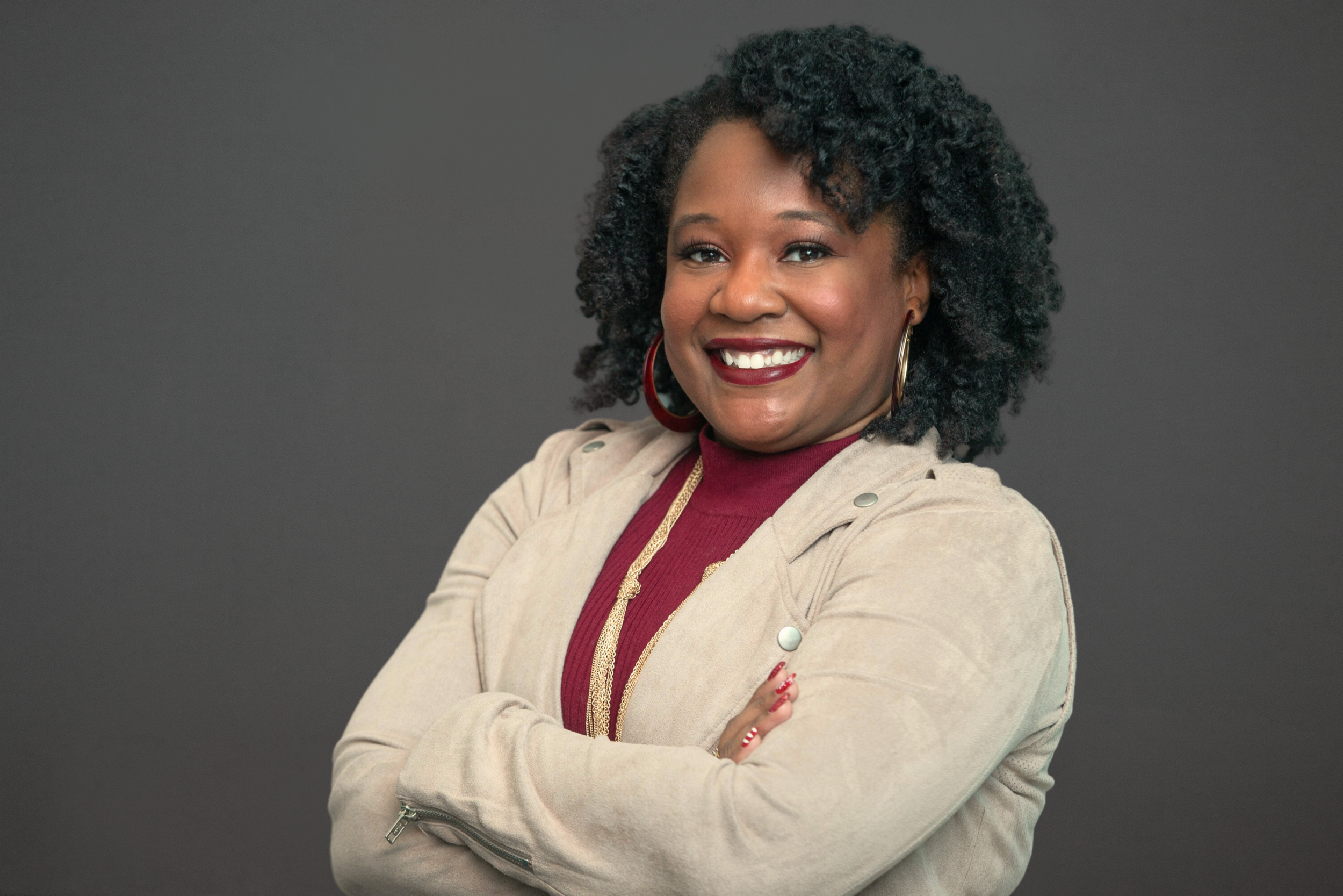 A woman with dark curly hair wearing a beige jacket and maroon top, smiling with crossed arms against a dark gray background.