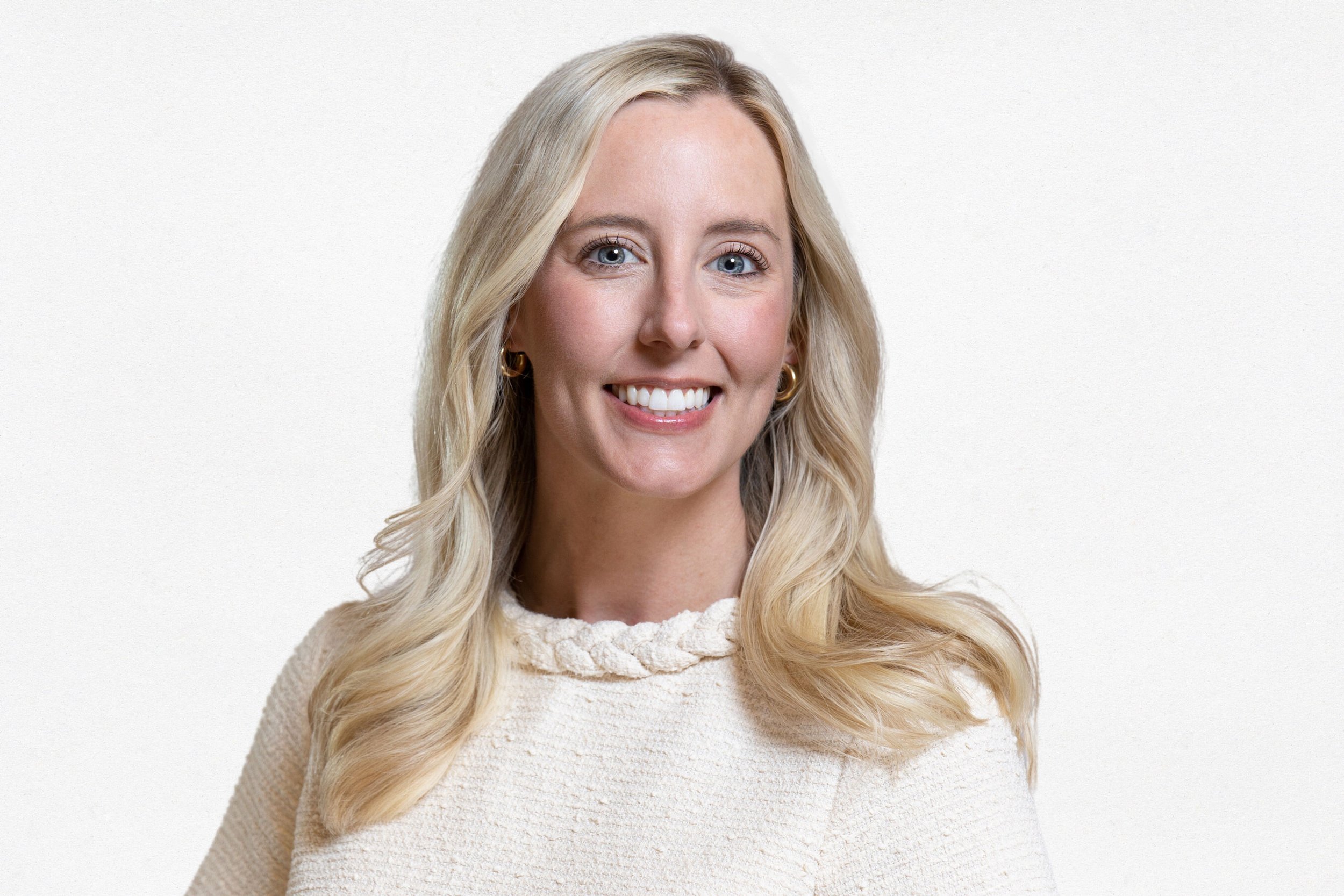 A woman with blonde hair smiling, wearing a cream-colored sweater and gold earrings, against a plain white background.