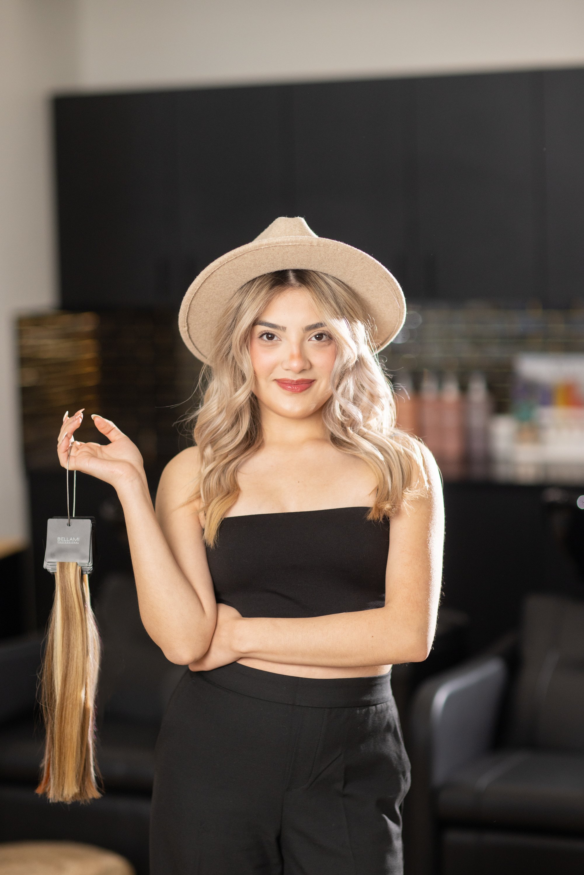 Young woman with blonde wavy hair wearing a beige hat and black top, holding a bundle of blonde hair extensions in her hand, smiling in a salon or studio.