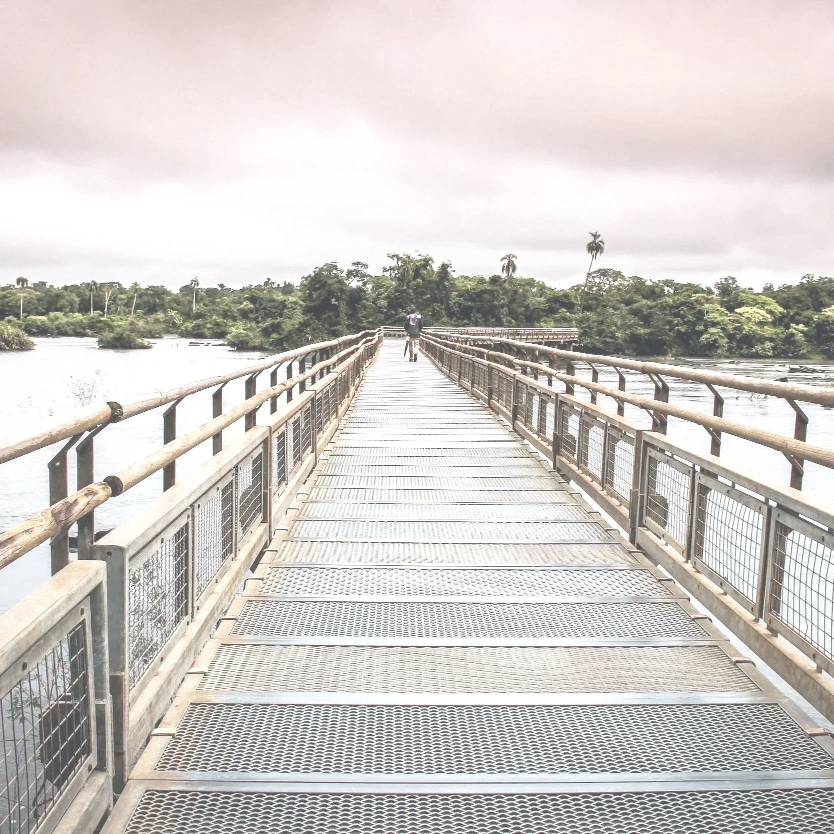A person walking on a metal bridge over a river, surrounded by dense forest and dark, cloudy sky.