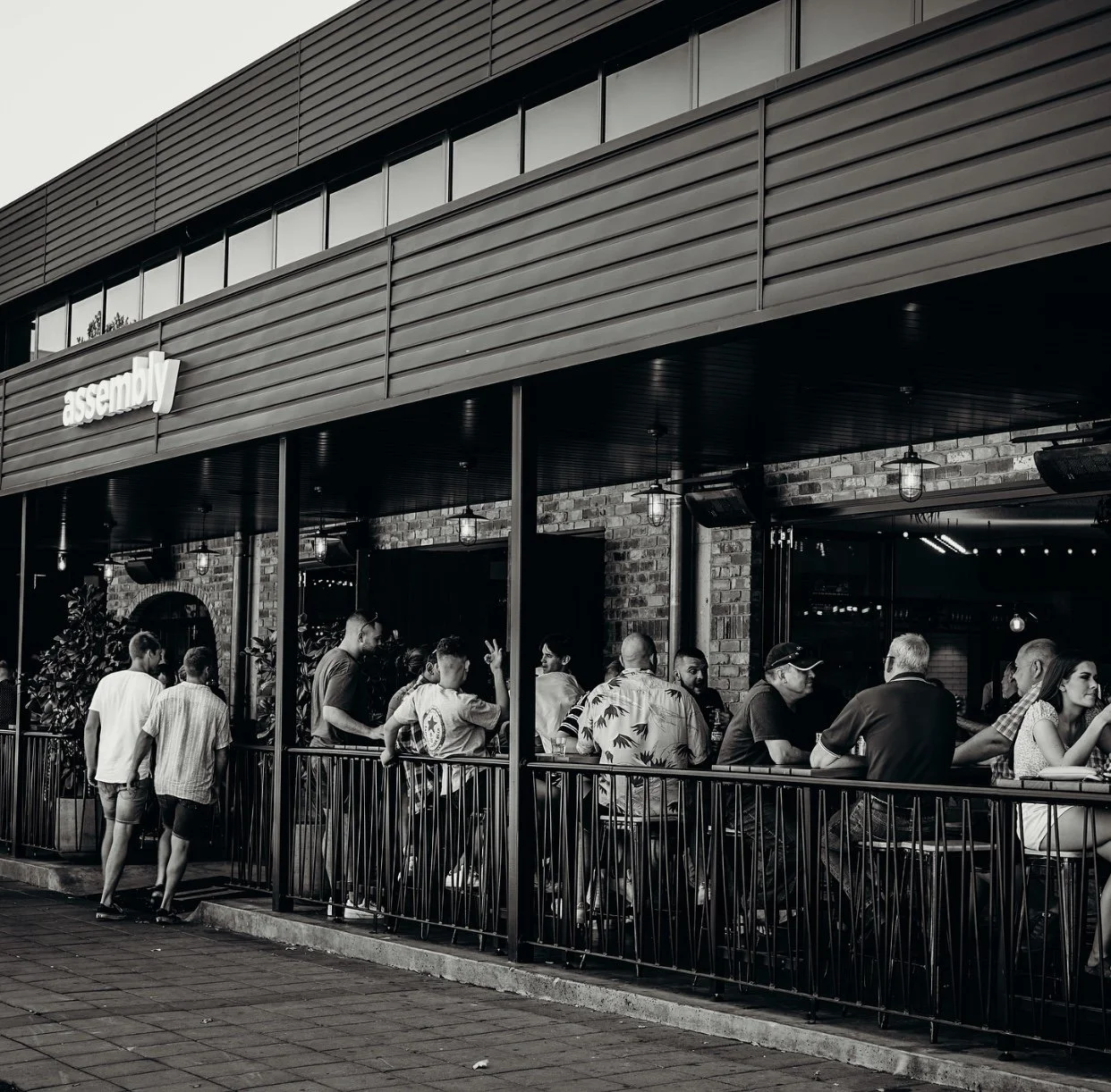 People sitting at a table on a restaurant patio, surrounded by brick walls and potted plants, with overhead lighting.