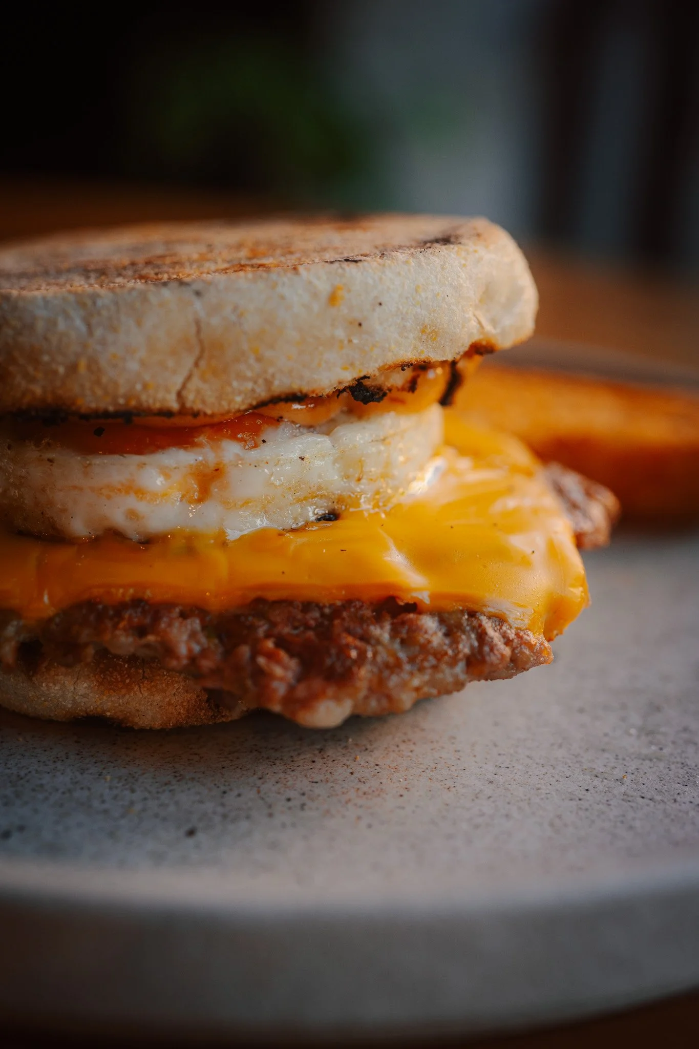 Close-up of a breakfast sandwich with a fried egg, melted cheddar cheese, sausage patty, and toasted bread on a plate.