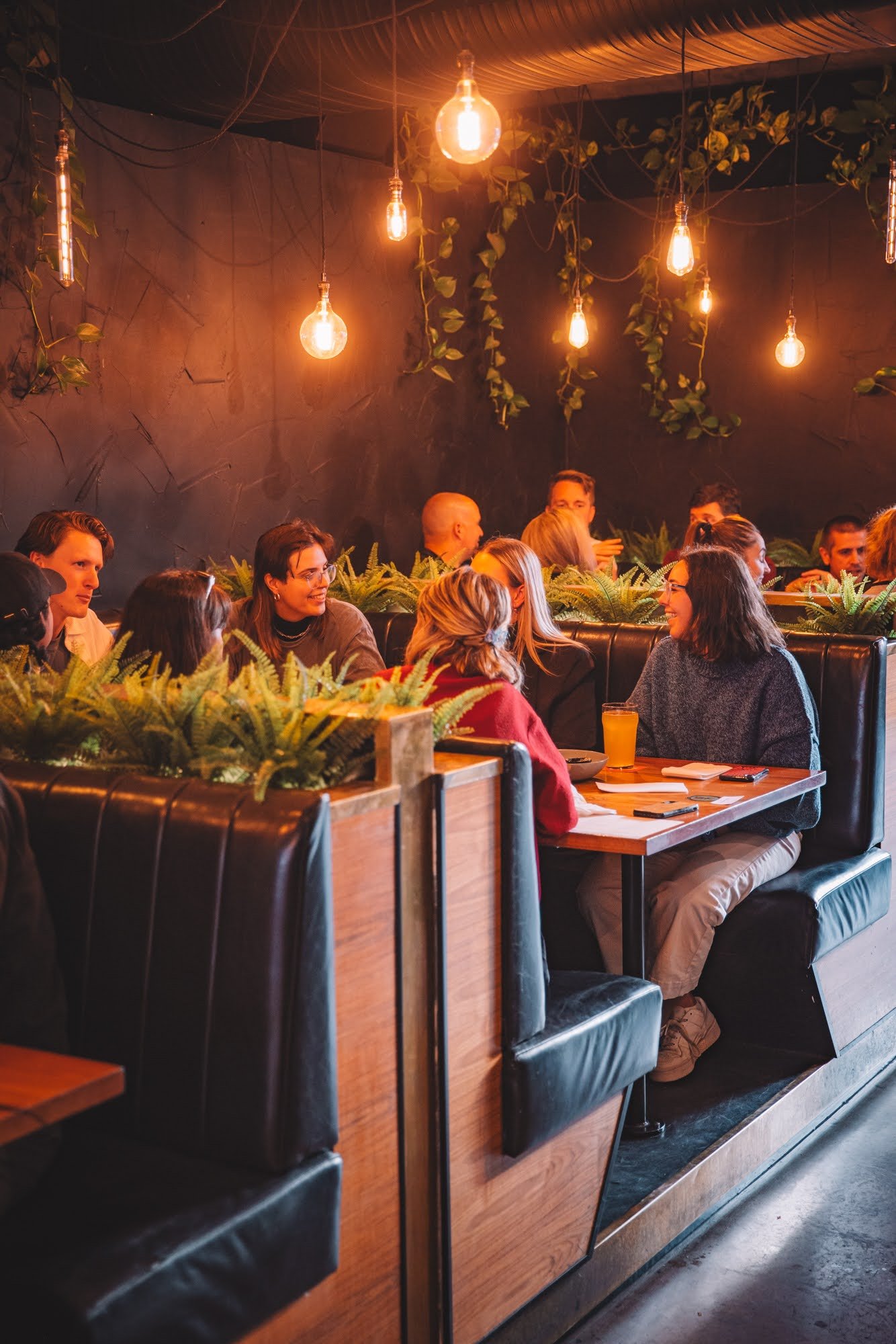 Group of people sitting in a restaurant booth under warm lighting and surrounded by plants.