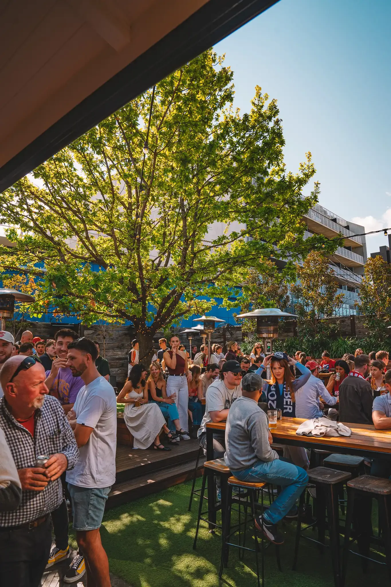 Outdoor patio with people sitting at tables under colorful umbrellas, trees in the background, and a musician playing guitar.