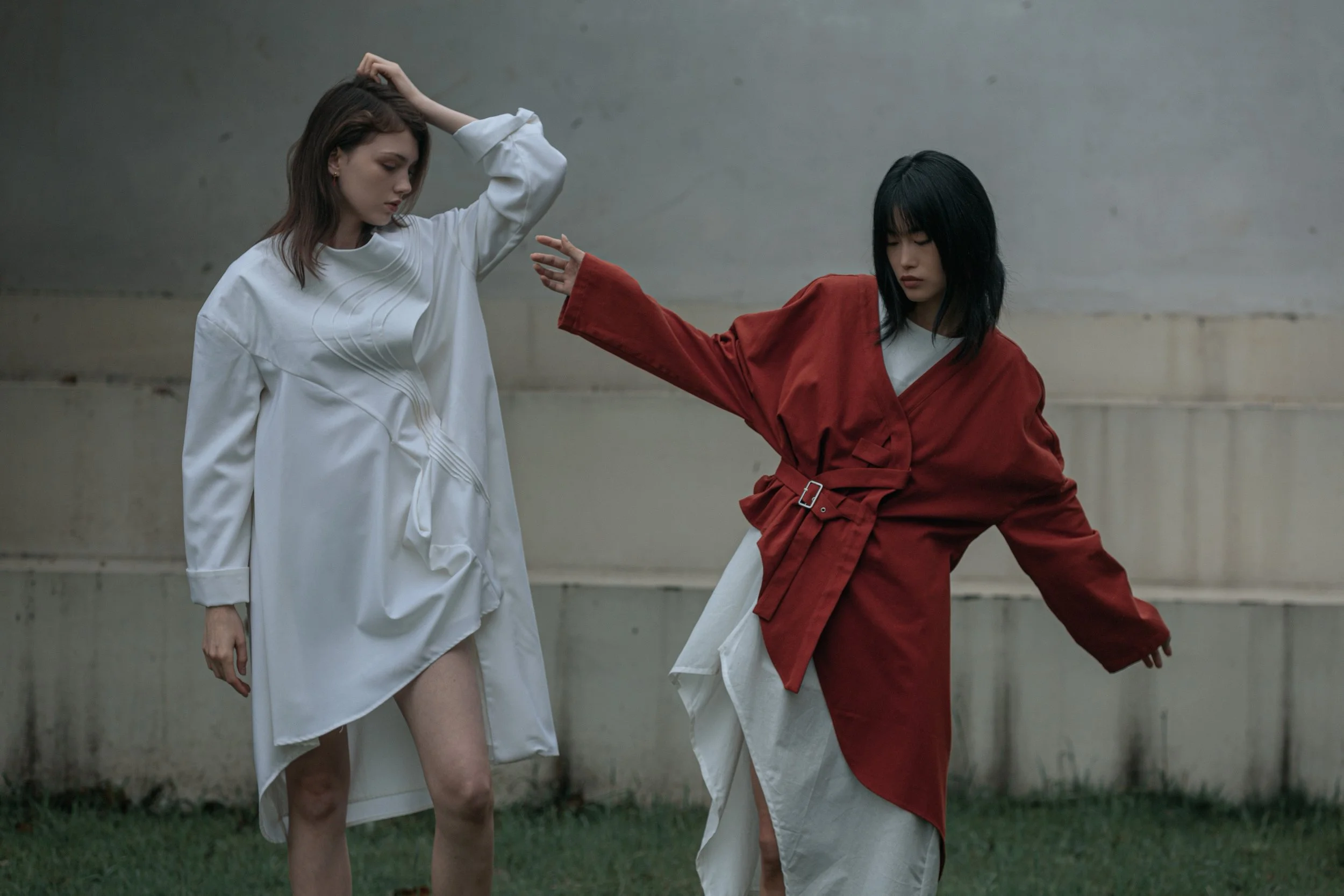 Two women dressed in stylish, loose-fitting dresses and jackets, posing outdoors against a concrete wall and grass, with one woman in a white dress and the other in a red jacket, with one hand raised and the other extended.