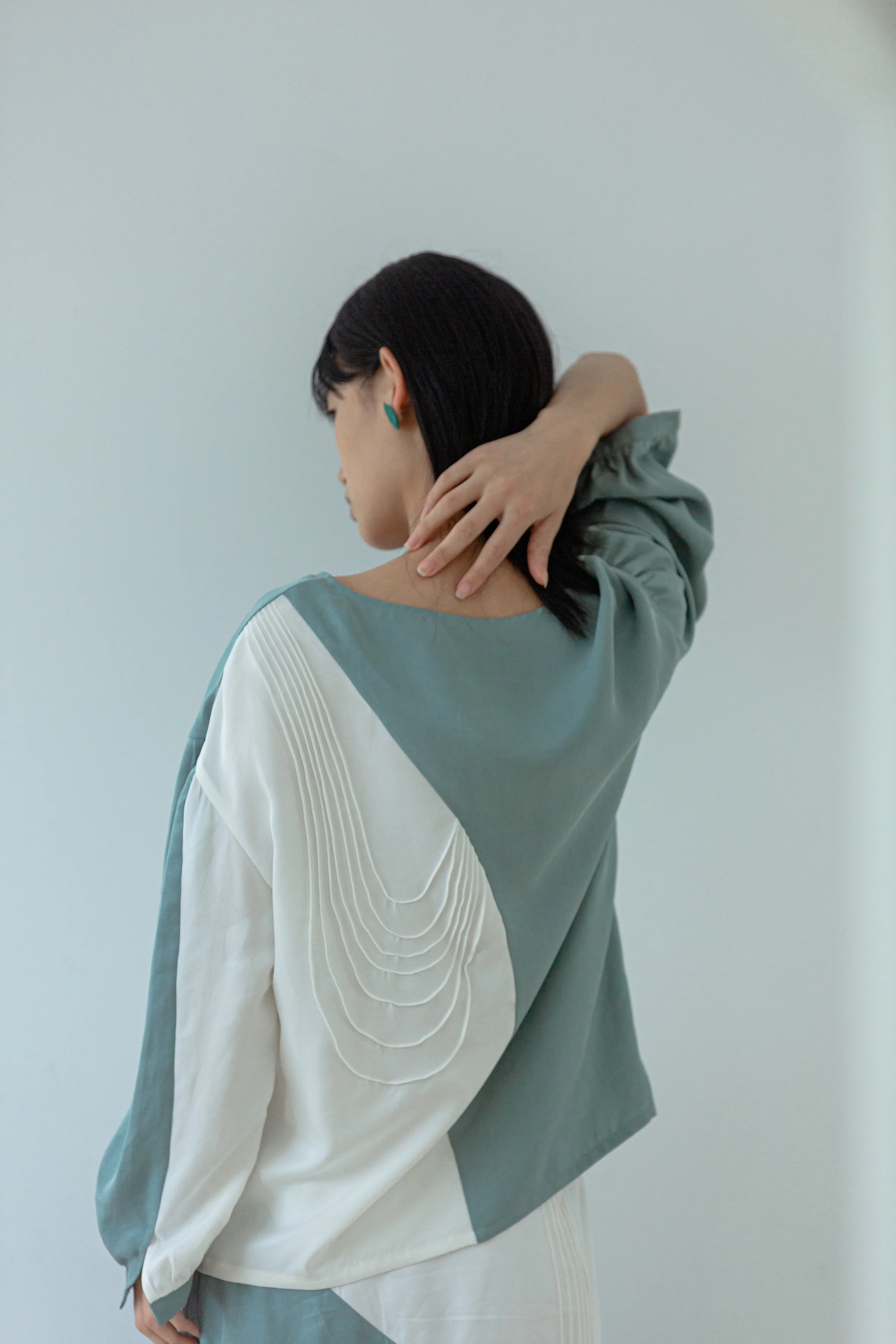 A woman with black hair, wearing teal earrings and a two-tone jacket with white and gray sections, stands with her back to the camera, touching her neck and looking downward against a plain, light-colored background.