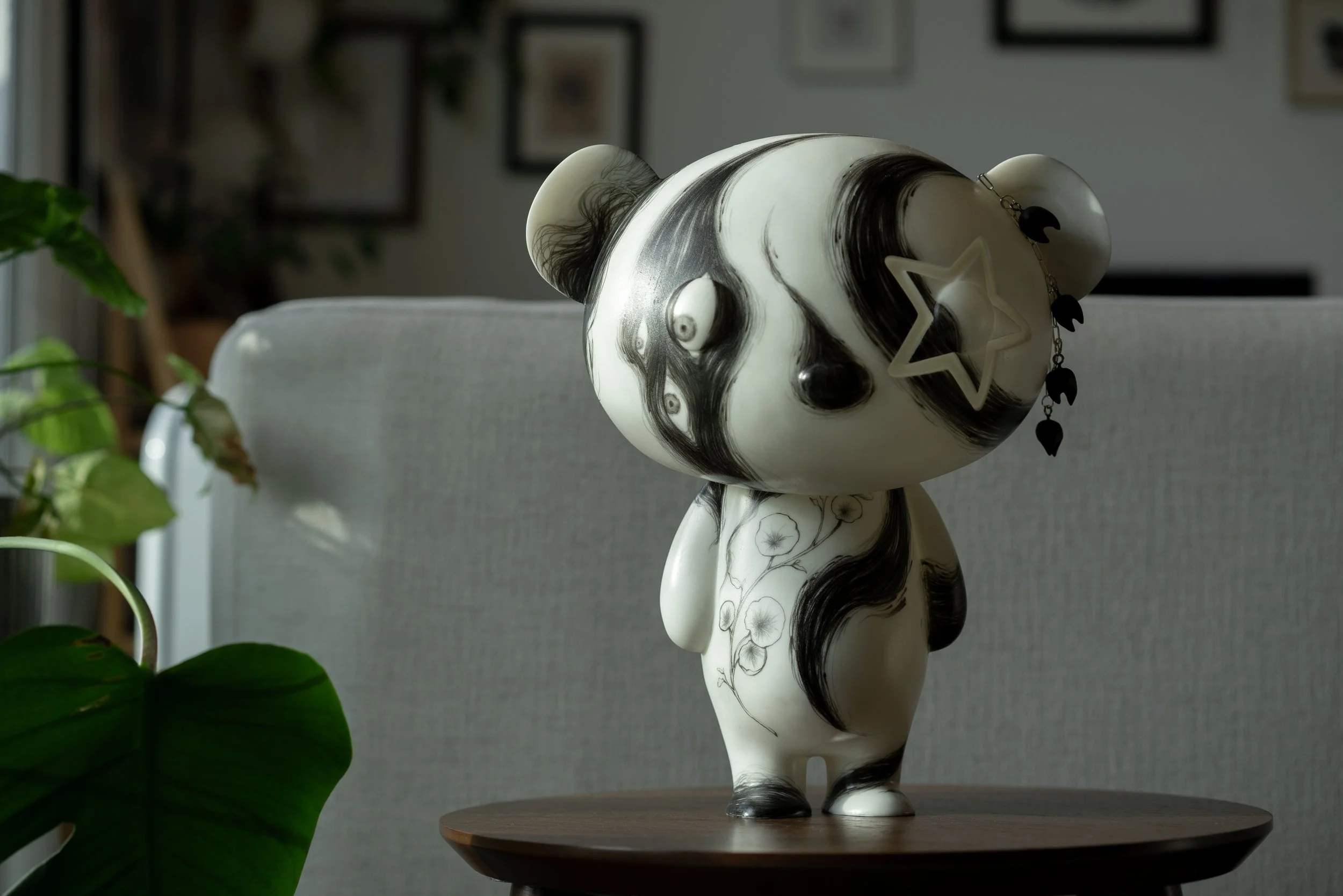 Decorative anthropomorphic figurine with a panda design, featuring black and white swirls and floral patterns, adorned with a star-shaped accessory and hanging black shapes, placed on a wooden table in a room with framed pictures and plants.