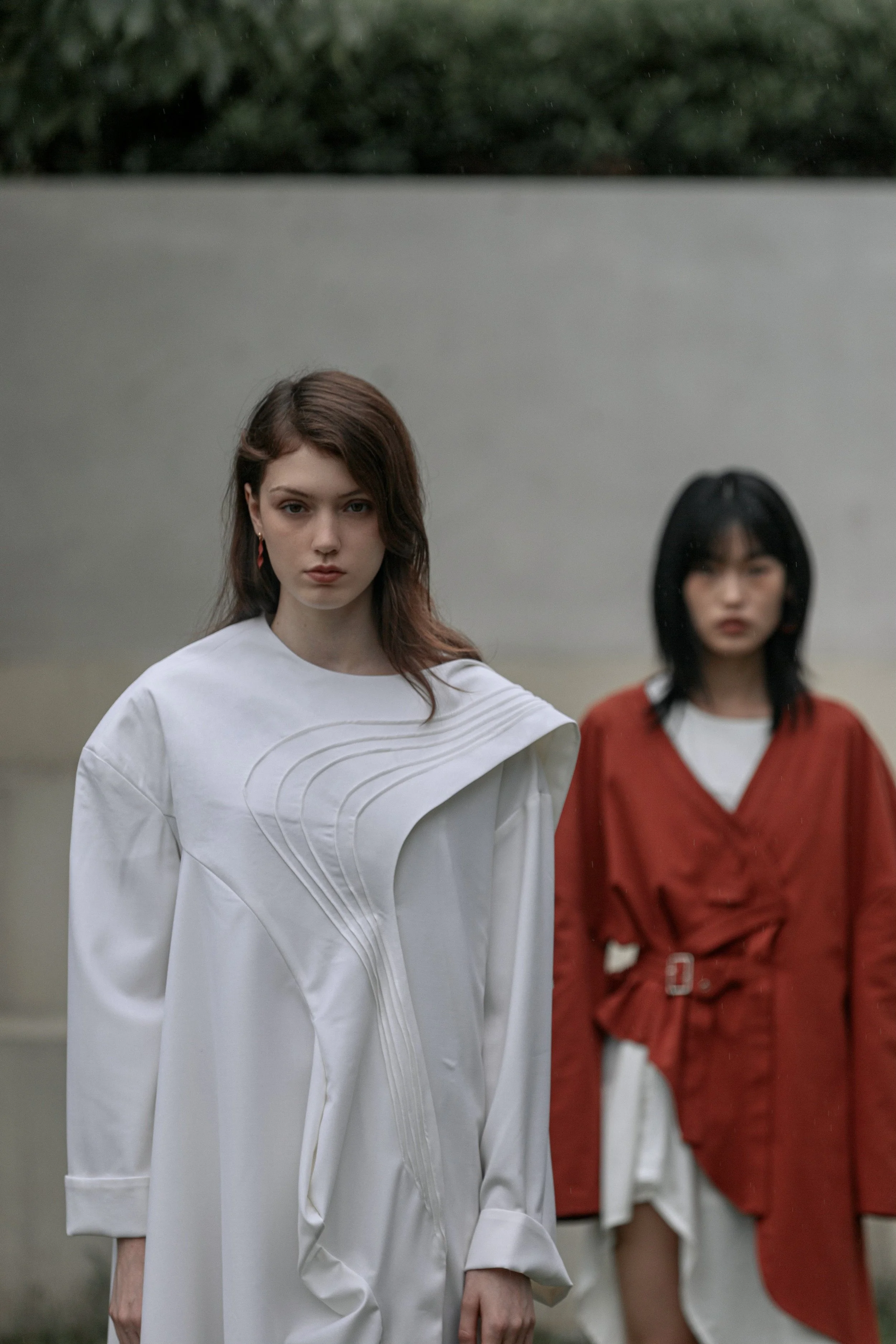 Two women standing outdoors in front of a concrete wall with greenery at the top. The woman in front has red hair, wears a white, duvet-like garment, and has a serious expression. The woman behind has black hair, wears a red jacket over a white shirt