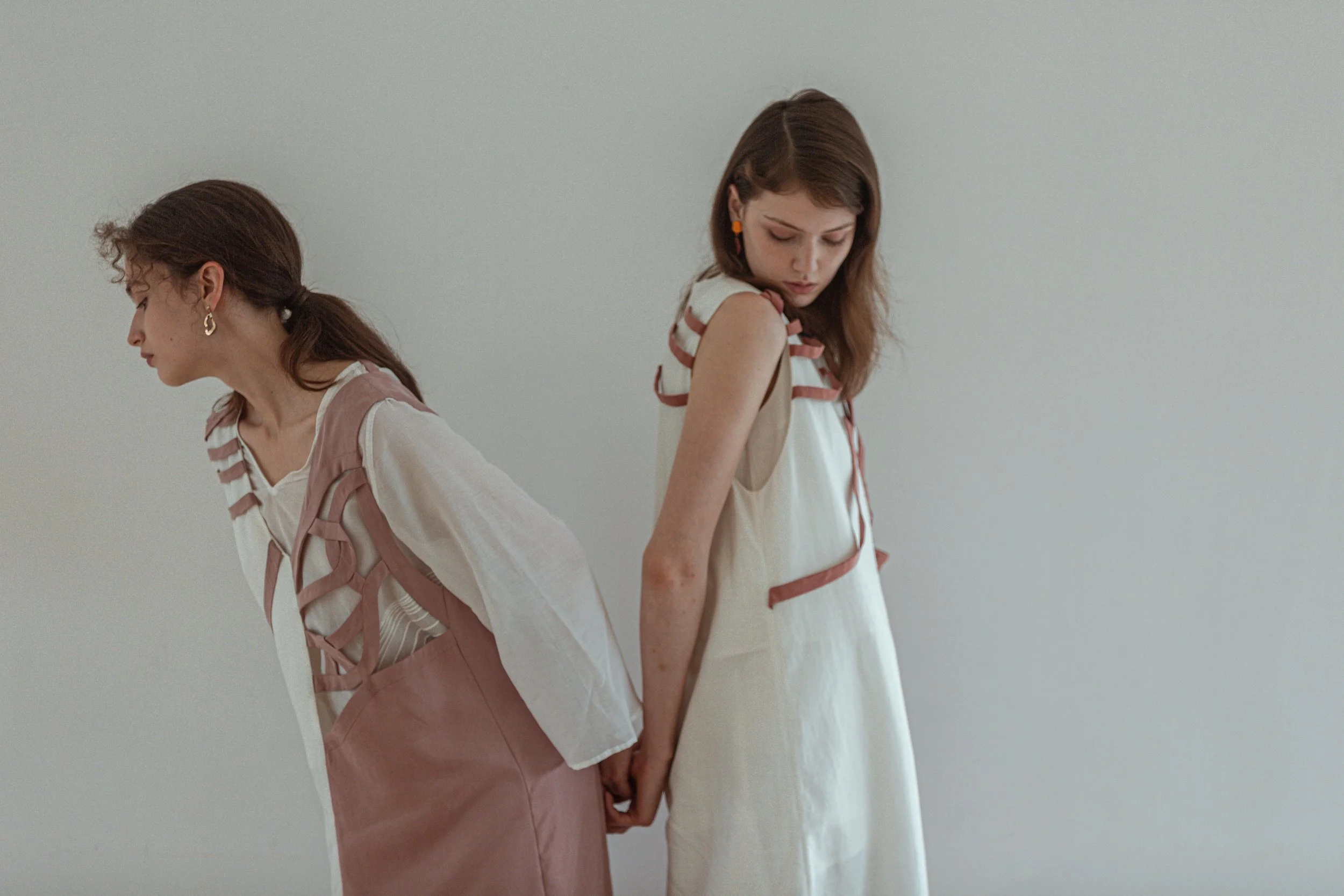 Two young women standing back to back, holding hands, wearing stylish outfits against a plain background.