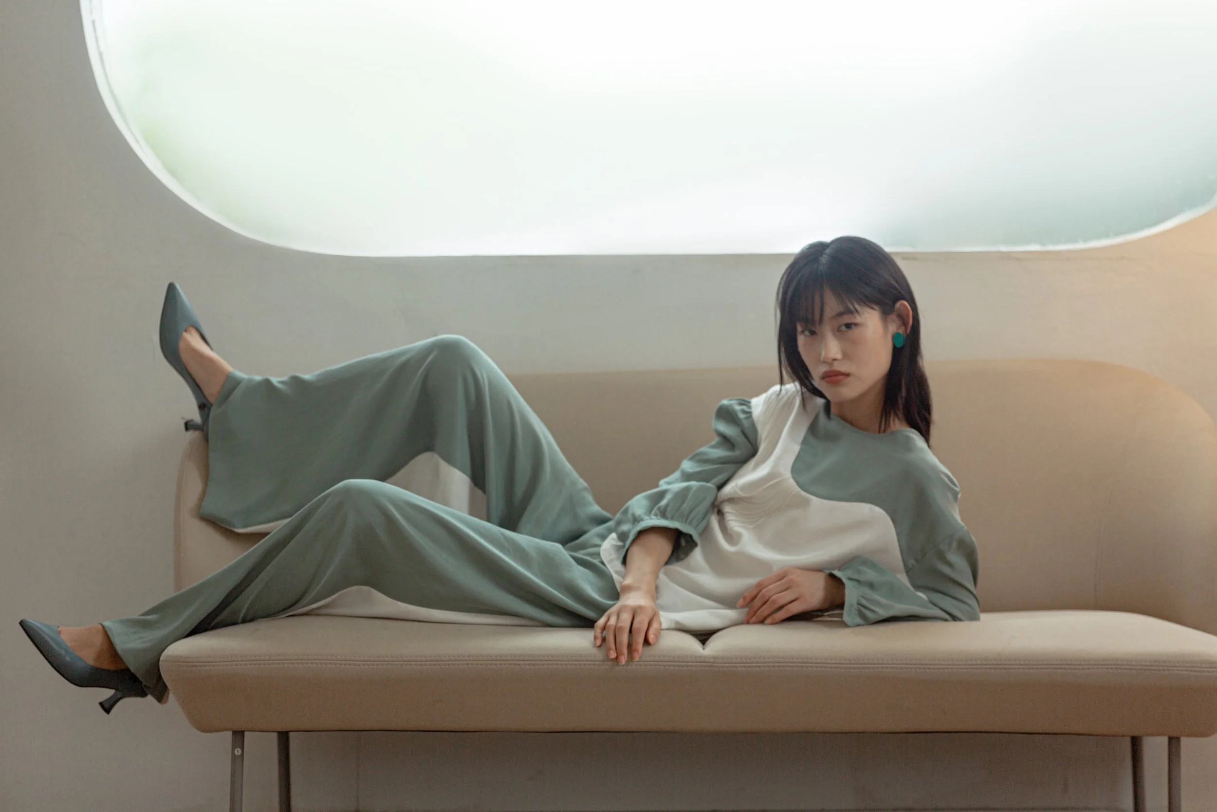 A woman with dark hair, wearing a stylish two-tone outfit with teal and white colors, and teal earrings, is lying on a beige modern sofa with her legs raised and crossed at the ankles. She is posing with a neutral expression in a minimalistic, softly