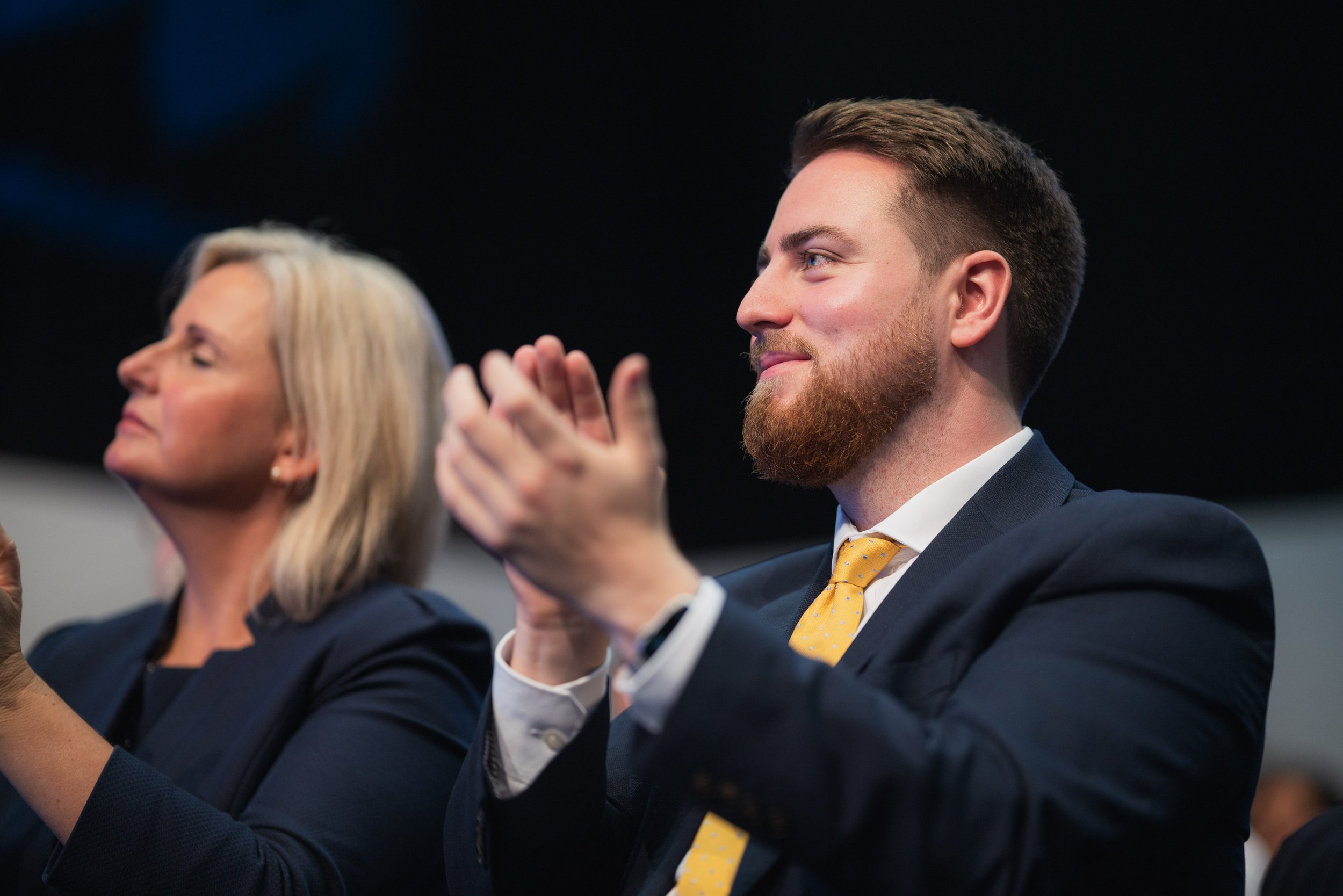 A man with a beard and a yellow tie clapping, sitting next to a woman with blonde hair during an event.