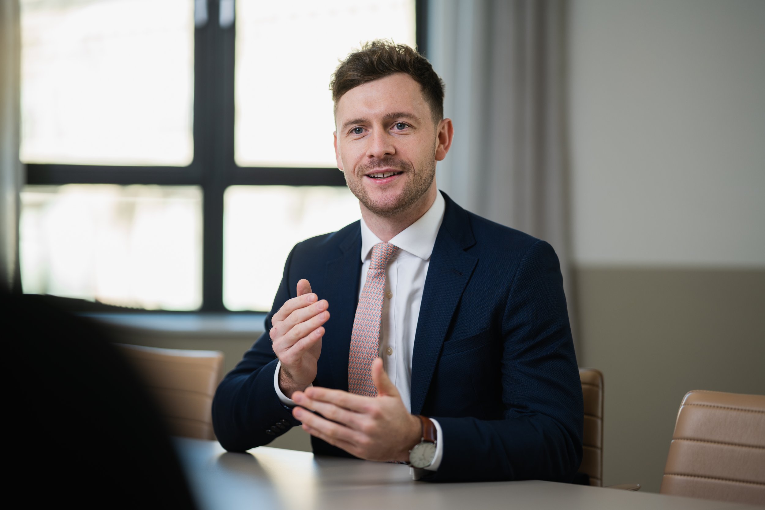Man in a suit speaking at a table, gesturing with hands, in an office setting.