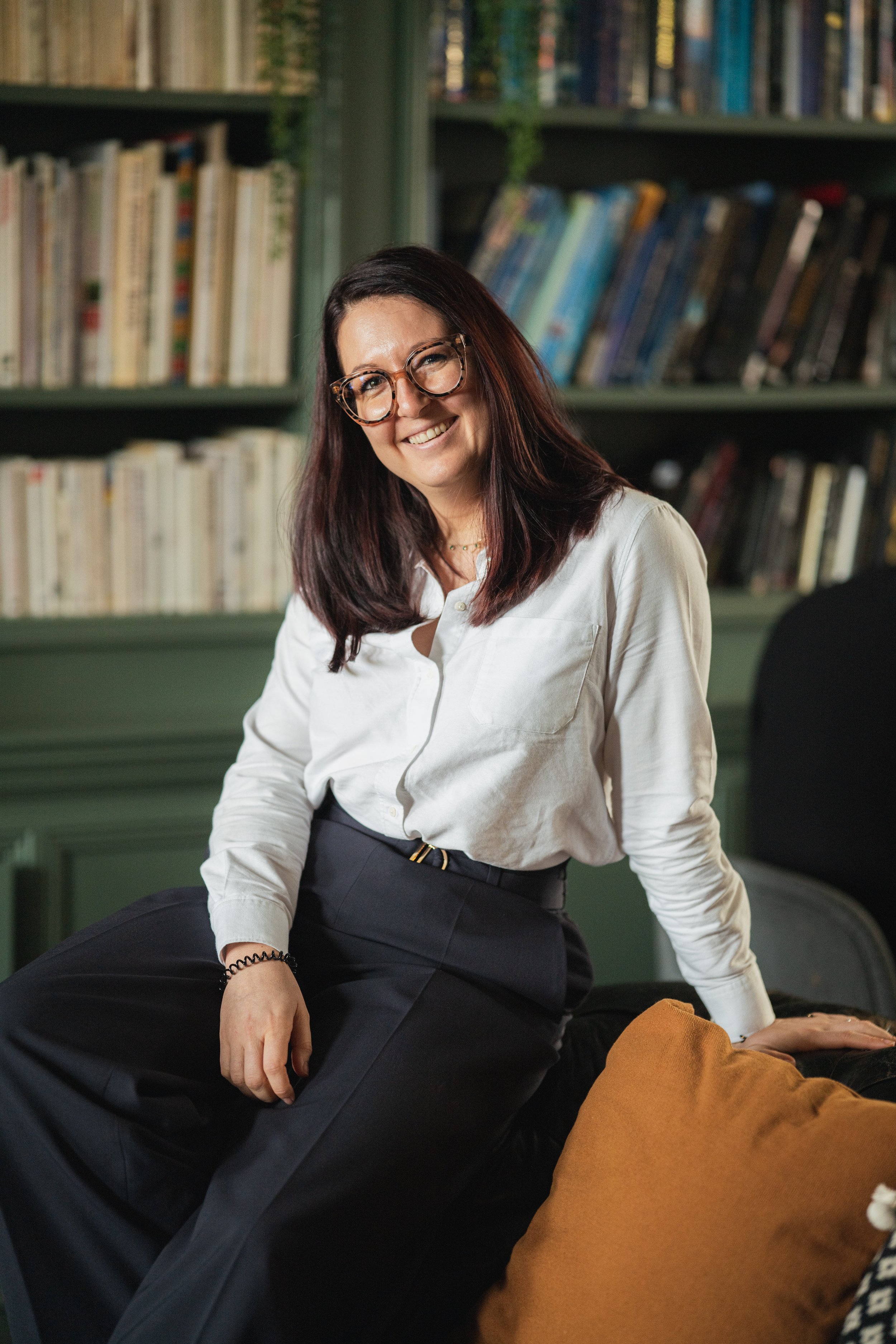 Candid business portrait of a woman with glasses sitting in front of bookshelves.