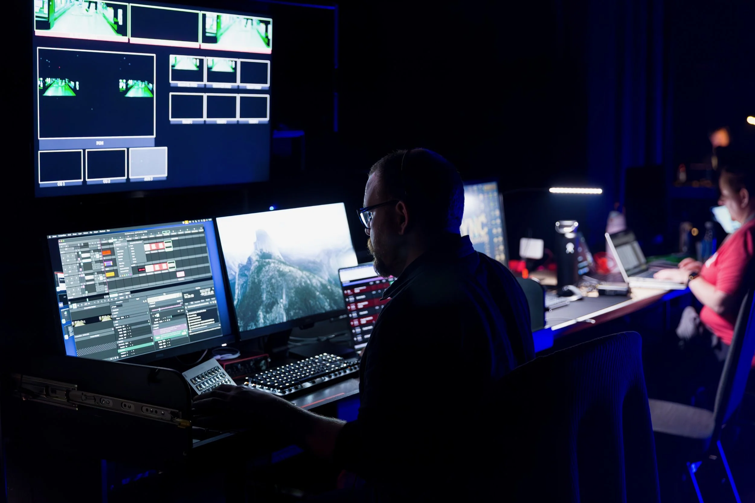 Two people working at multiple computer monitors in a dark room, with one large screen displaying a grid of video feeds and the others showing music production software and a mountain landscape wallpaper.