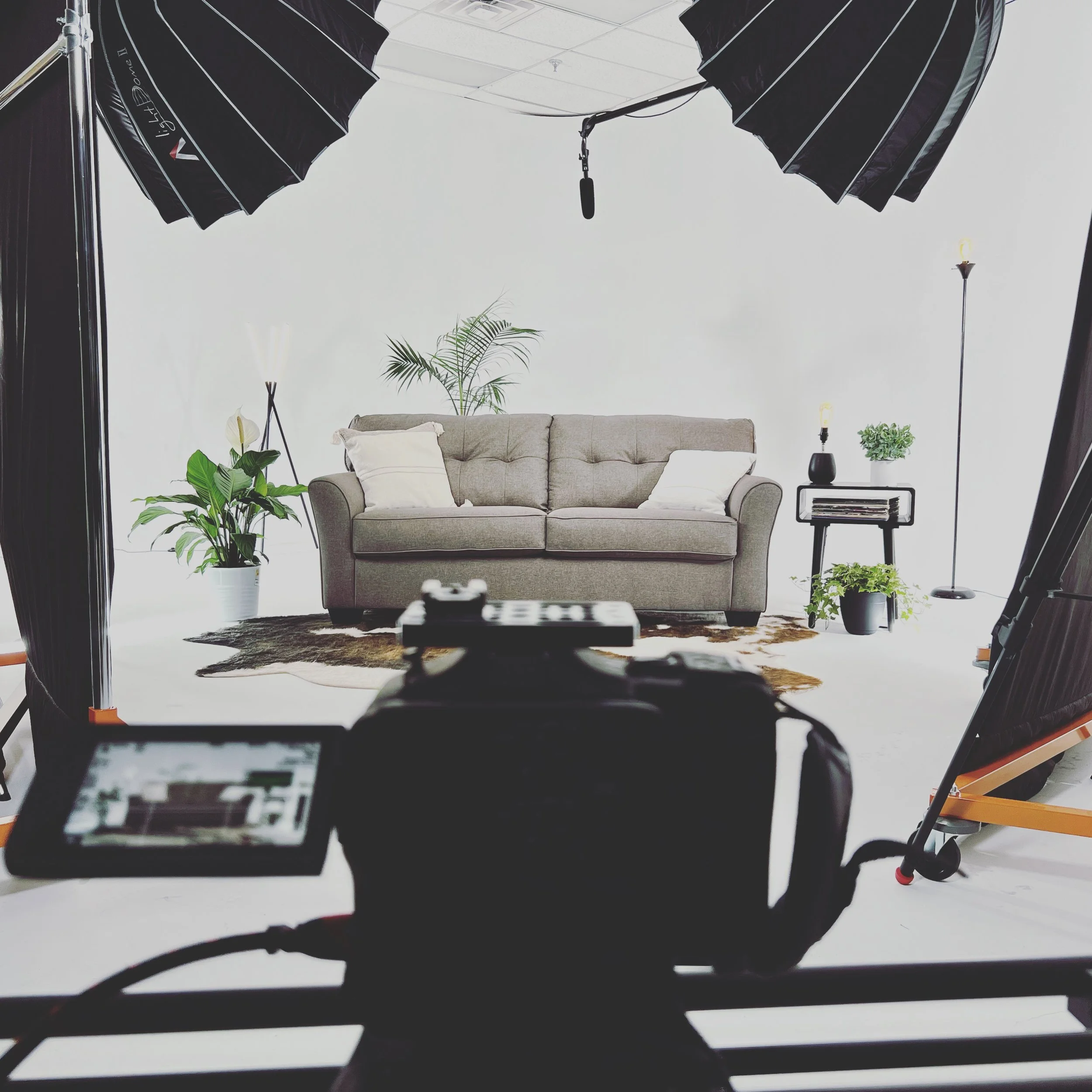 Photo of a professional video shoot setup showing a sofa with pillows, plants, and side table in a studio with lighting equipment and camera in the foreground.