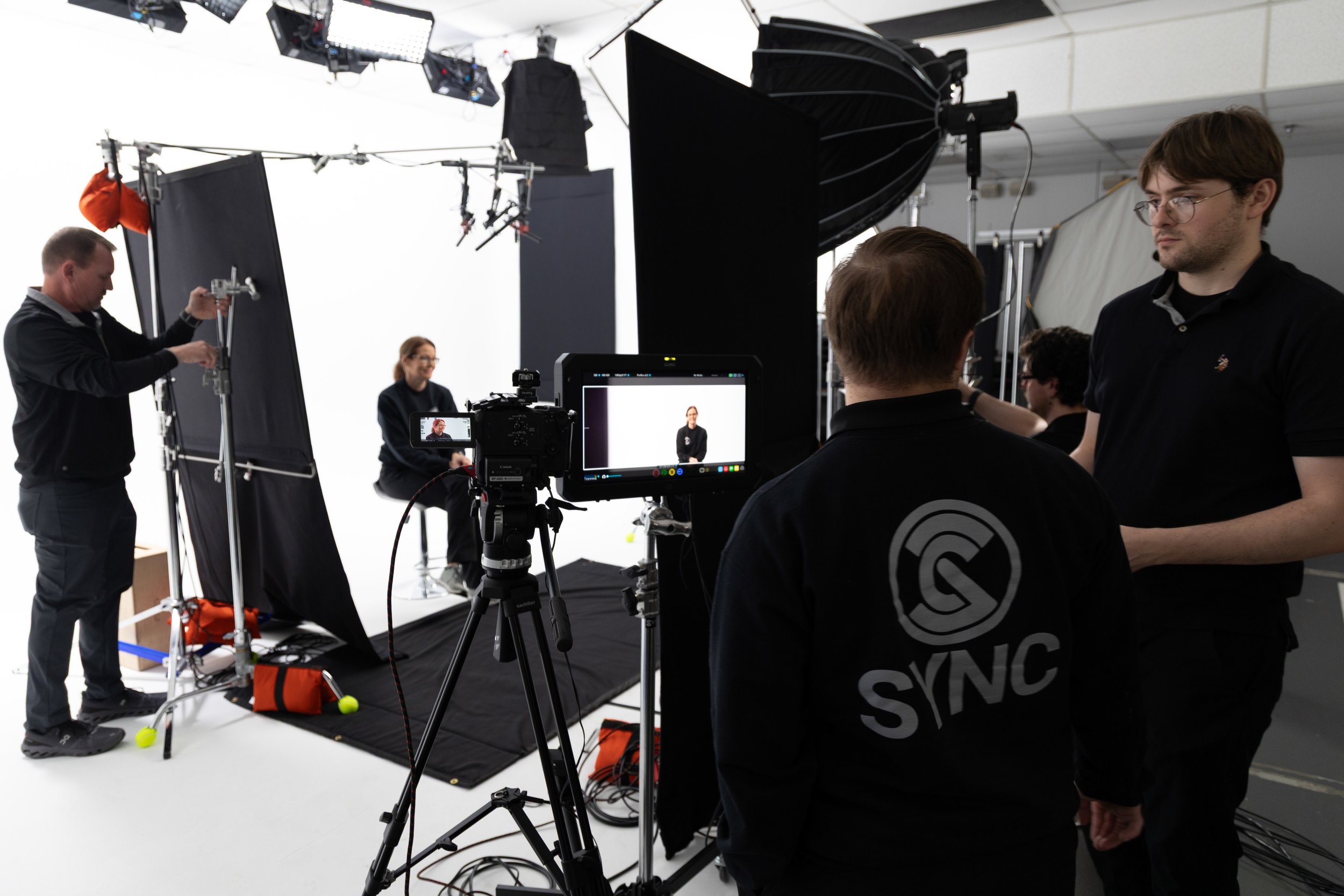 Behind the scenes of a video shoot at a studio with people setting up cameras and equipment in front of a green screen, including a woman seated and a woman speaking to camera.