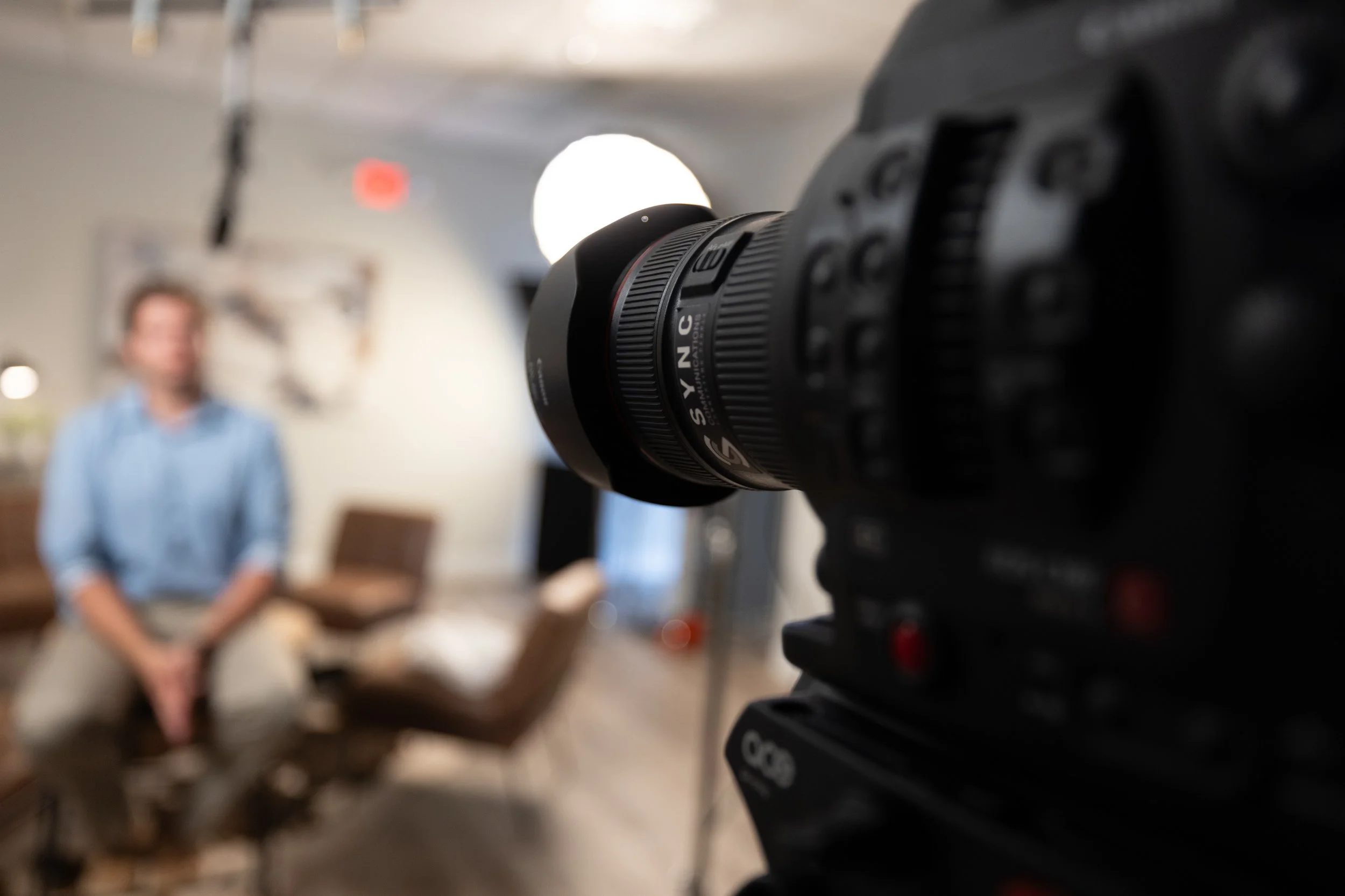 Close-up of a professional video camera focusing on a blurred person sitting in the background during an interview or recording session.