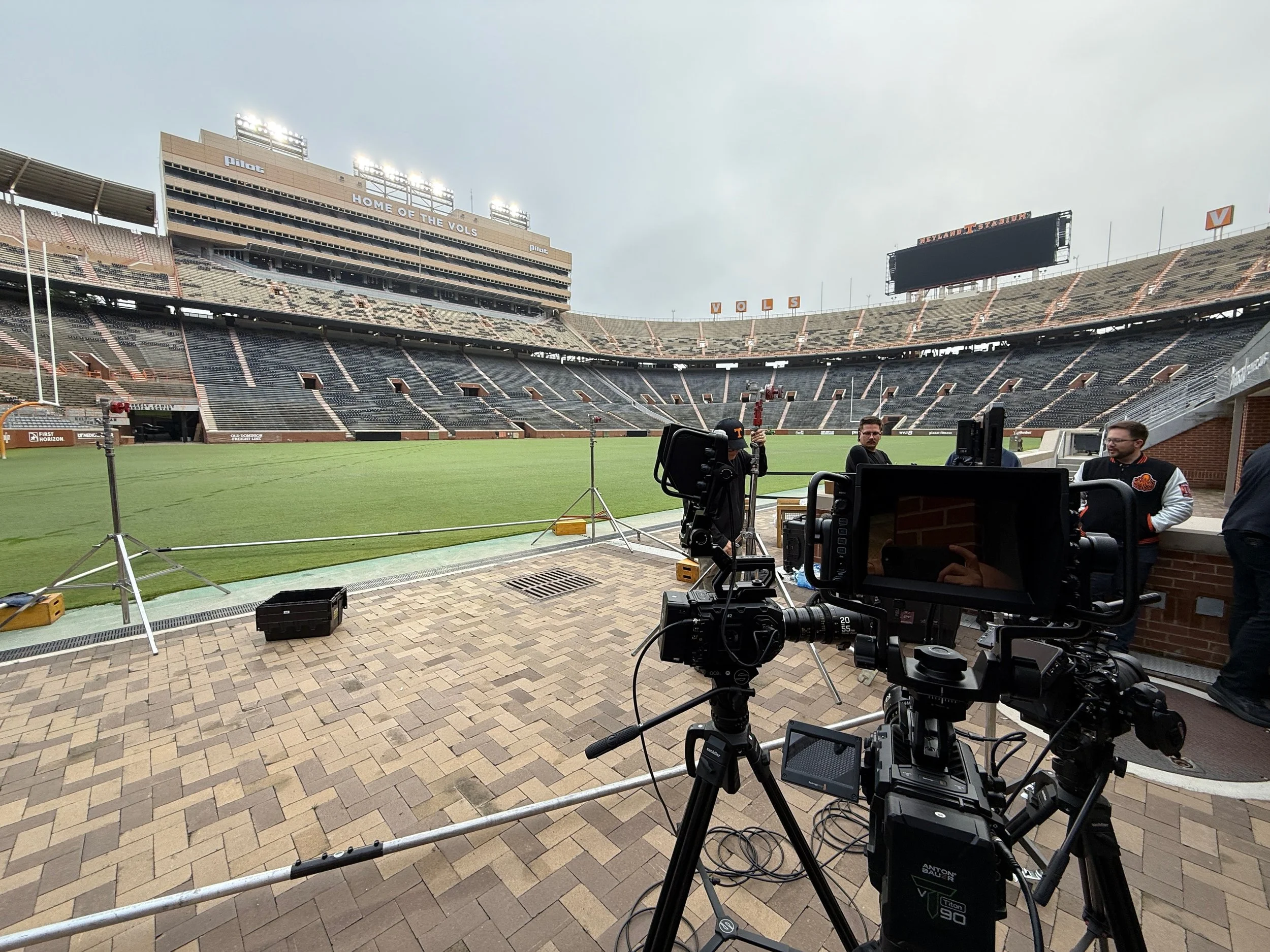View of an empty football stadium with camera equipment and crew setting up for filming or broadcasting, overcast sky above.