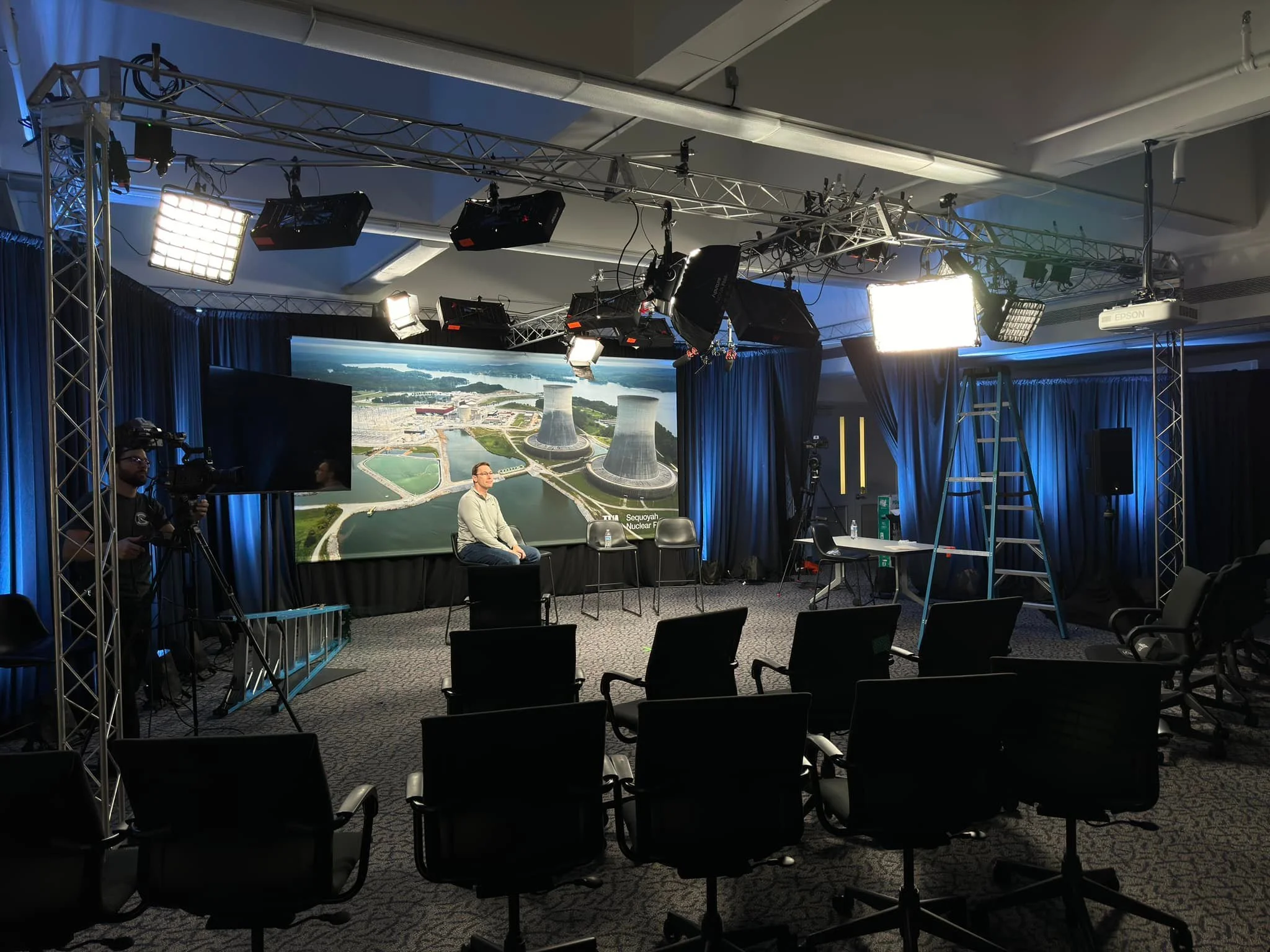 Recording studio with professional lighting, large screen displaying an aerial view of a nuclear power plant, a woman seated in front of the screen, and camera crew setting up equipment.