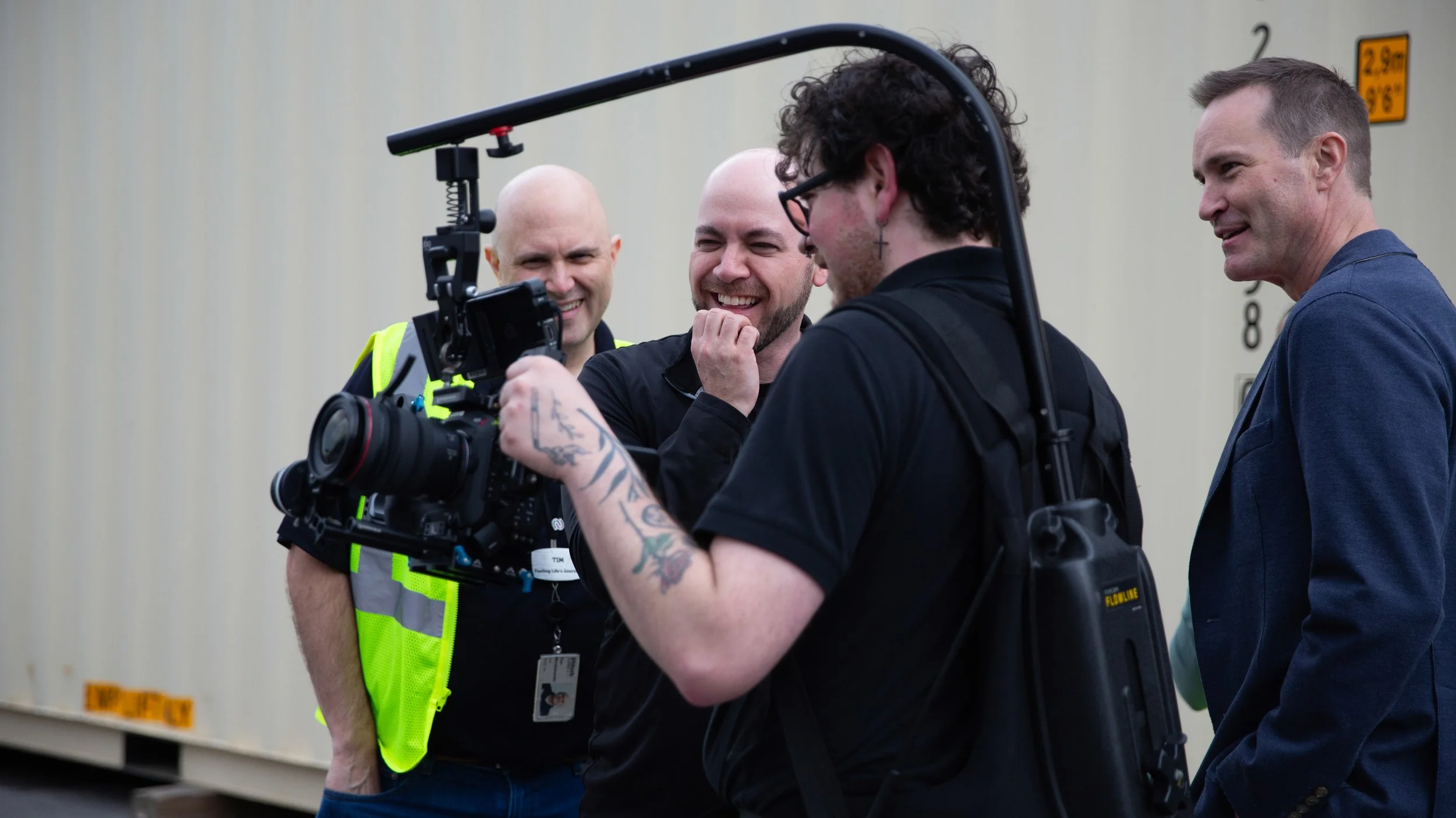 Four men are gathered outdoors, looking at a camera held by one of them, all smiling and laughing, near a beige building with height markers.
