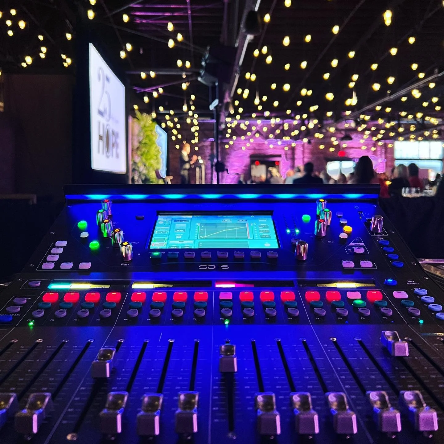 A professional audio mixing console with colorful illuminated controls in a dimly lit event space decorated with string lights and people in the background.