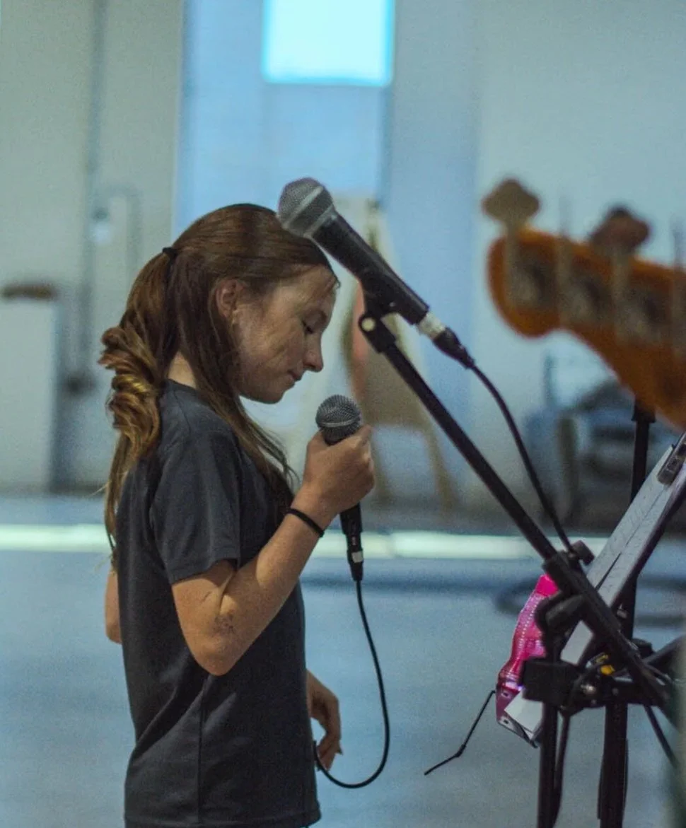 A young girl with red hair in a braid, holding a microphone, standing in front of a microphone stand and a music stand, in a studio or rehearsal space.