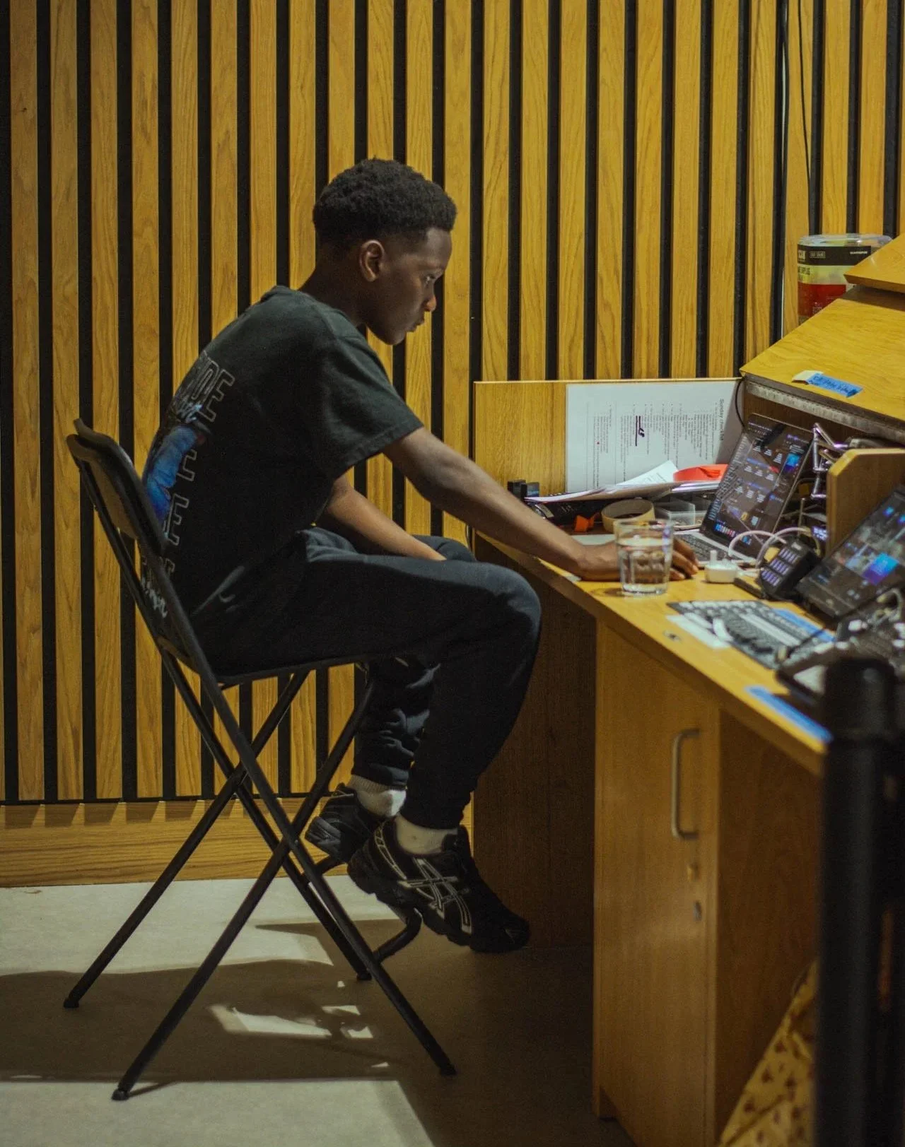 A young man sitting at a wooden desk with multiple electronic devices, including a laptop and two phones, in a room with wood-paneled walls. He's wearing a black graphic t-shirt, black pants, and black sneakers, with a glass of water nearby.