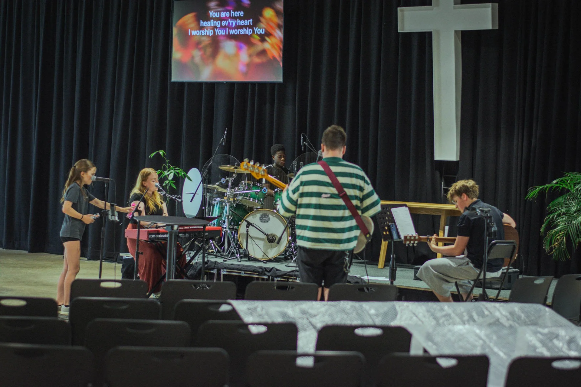 A stage with a black curtain and white cross, where a group of young musicians are rehearsing or performing. There are four performers: two girls with microphones and a keyboard, one boy playing guitar, and one boy on drums. A large screen above displays song lyrics, and there are plants on either side of the stage.