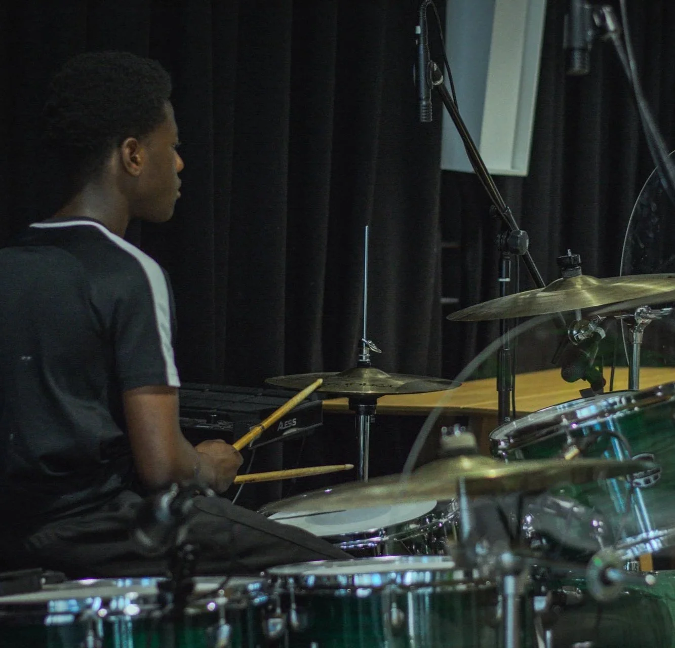 A person sitting at a drum set in a dark room, holding drumsticks, with black curtains and microphones around.