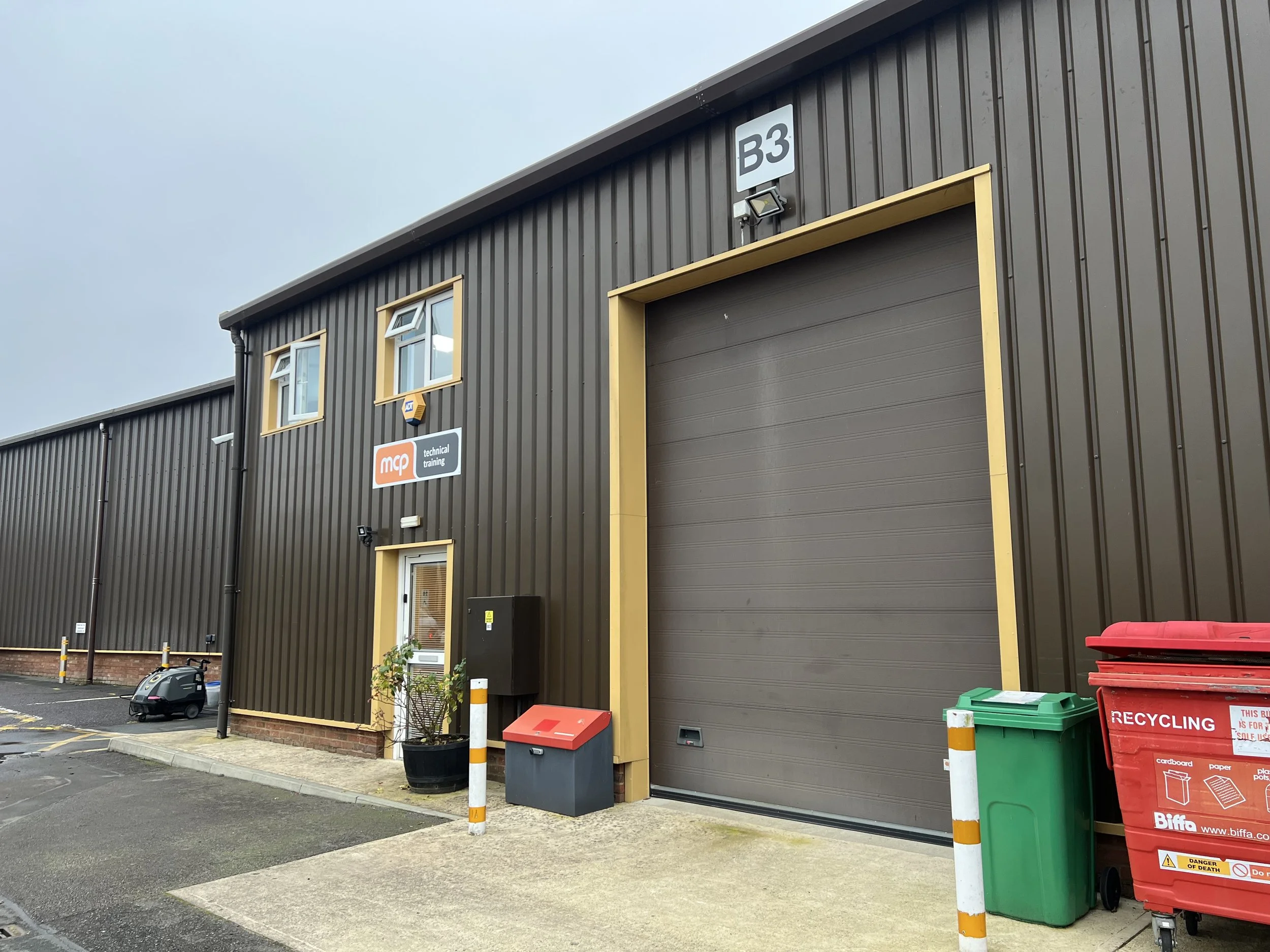 Exterior view of a brown industrial building with a large roller door, small windows, and a sign that says 'mcq technical training.' There are recycling bins and a small plant outside.