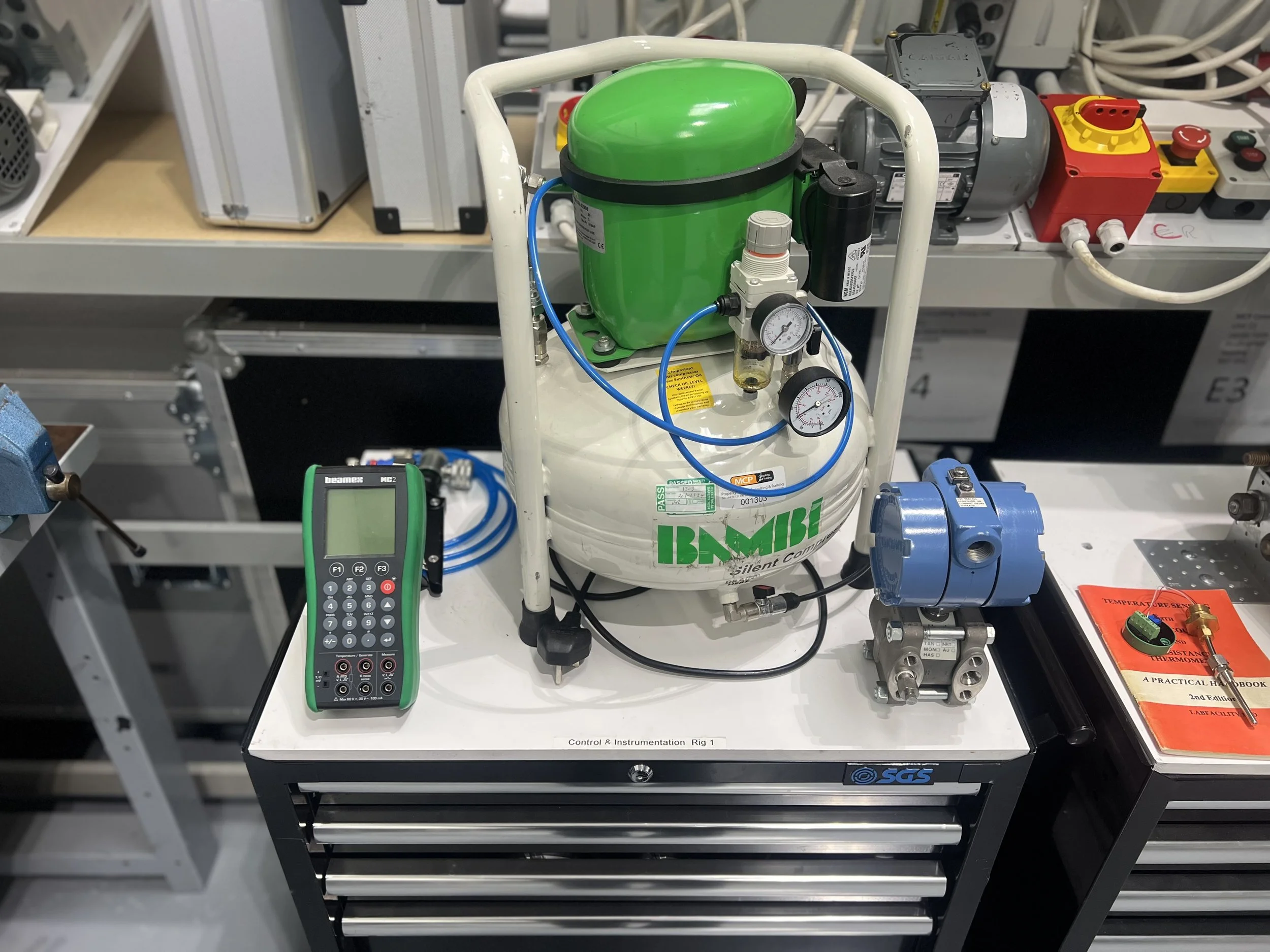Laboratory bench with a green and white ABAC portable air compressor, a digital calibration tool, and various technical components and gauges.