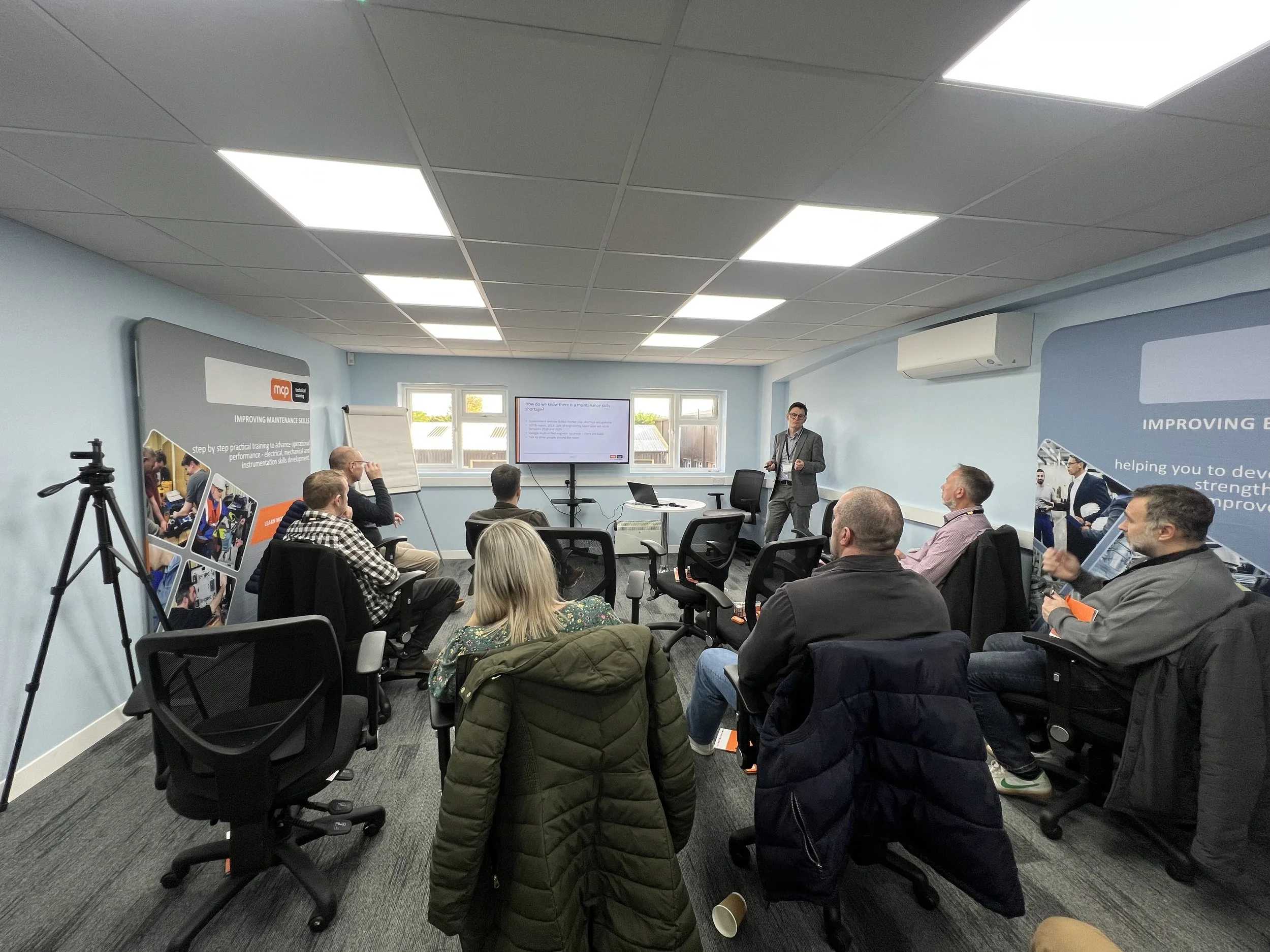 A man in a gray suit giving a presentation to a group of people seated in a conference room with light blue walls, a ceiling with fluorescent lights, and a large screen displaying a slide. Attendees are listening attentively, some taking notes, with
