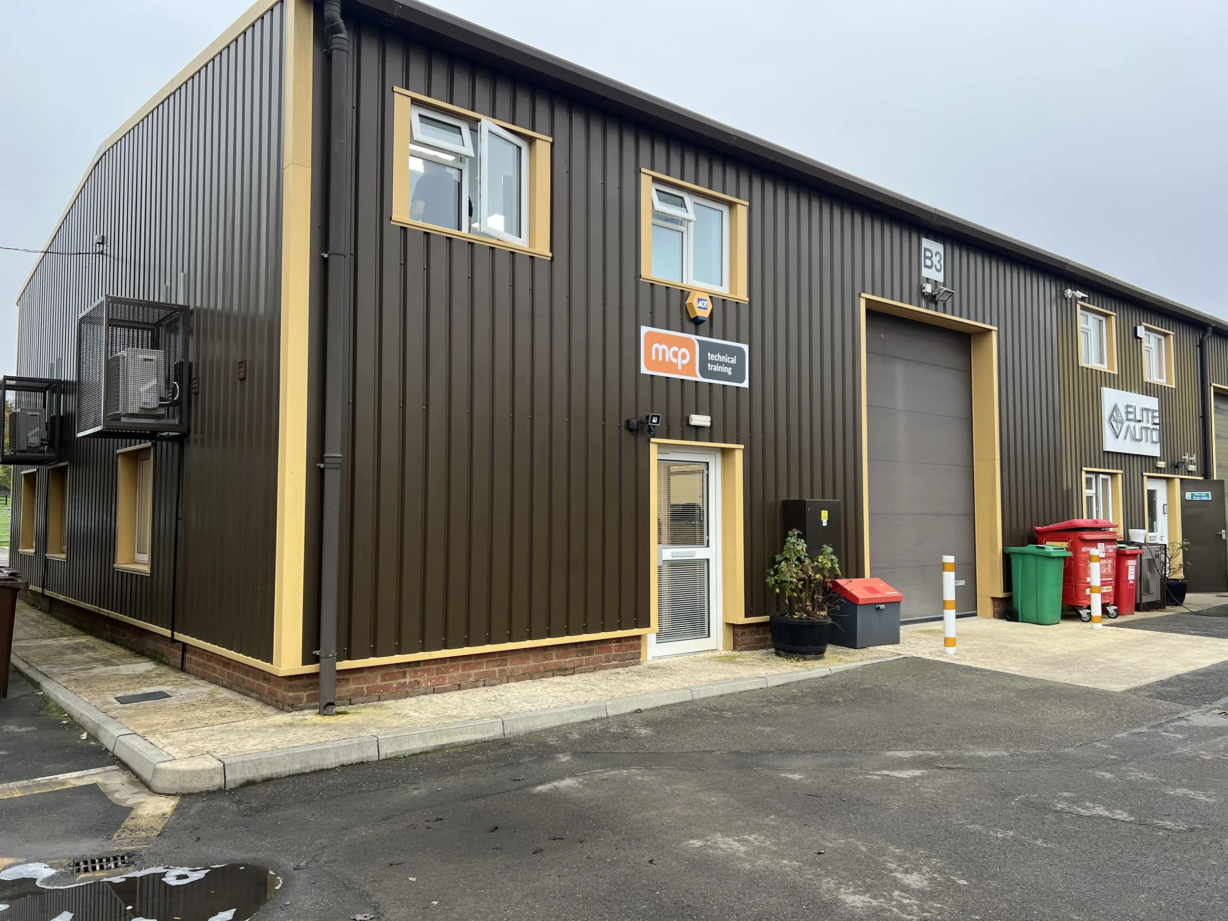 A modern industrial building with black metal exterior, yellow trim, and a large garage door, situated in a parking lot with trash bins and potted plants outside.