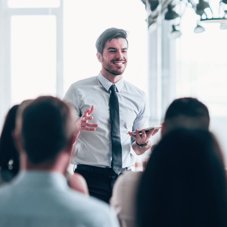 A man with dark hair, beard, wearing a white shirt and black tie, is smiling and speaking to a group of people in an office conference room.