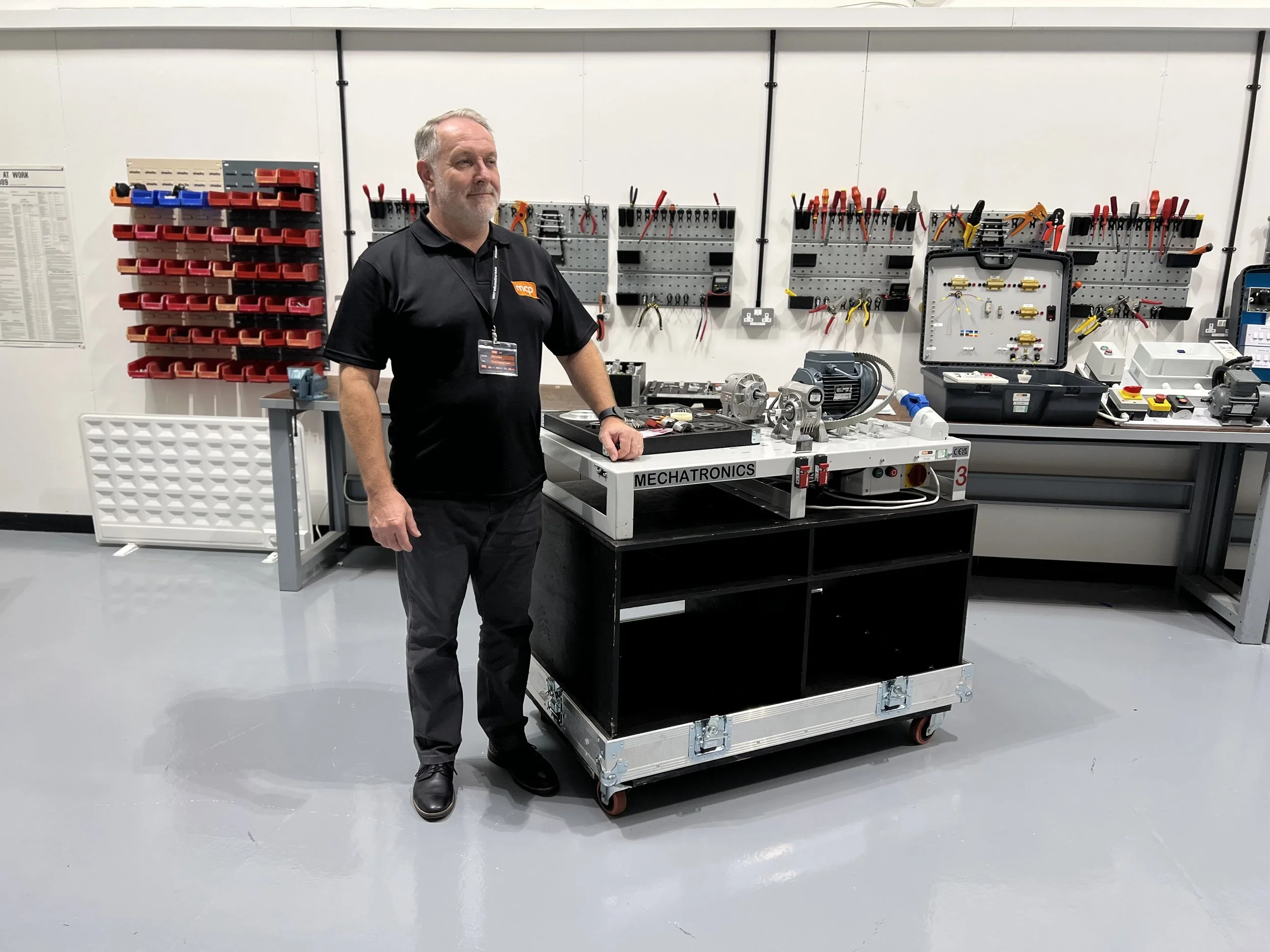 A man in black work attire standing beside a mechanical work station in a workshop, with tools and equipment in the background.