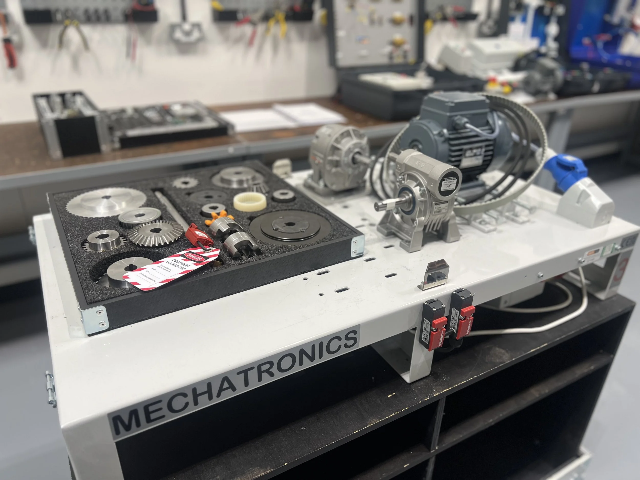 Workstation with various gears, pulleys, and mechanical parts on a white table labeled 'MECHATROICS' in a workshop.