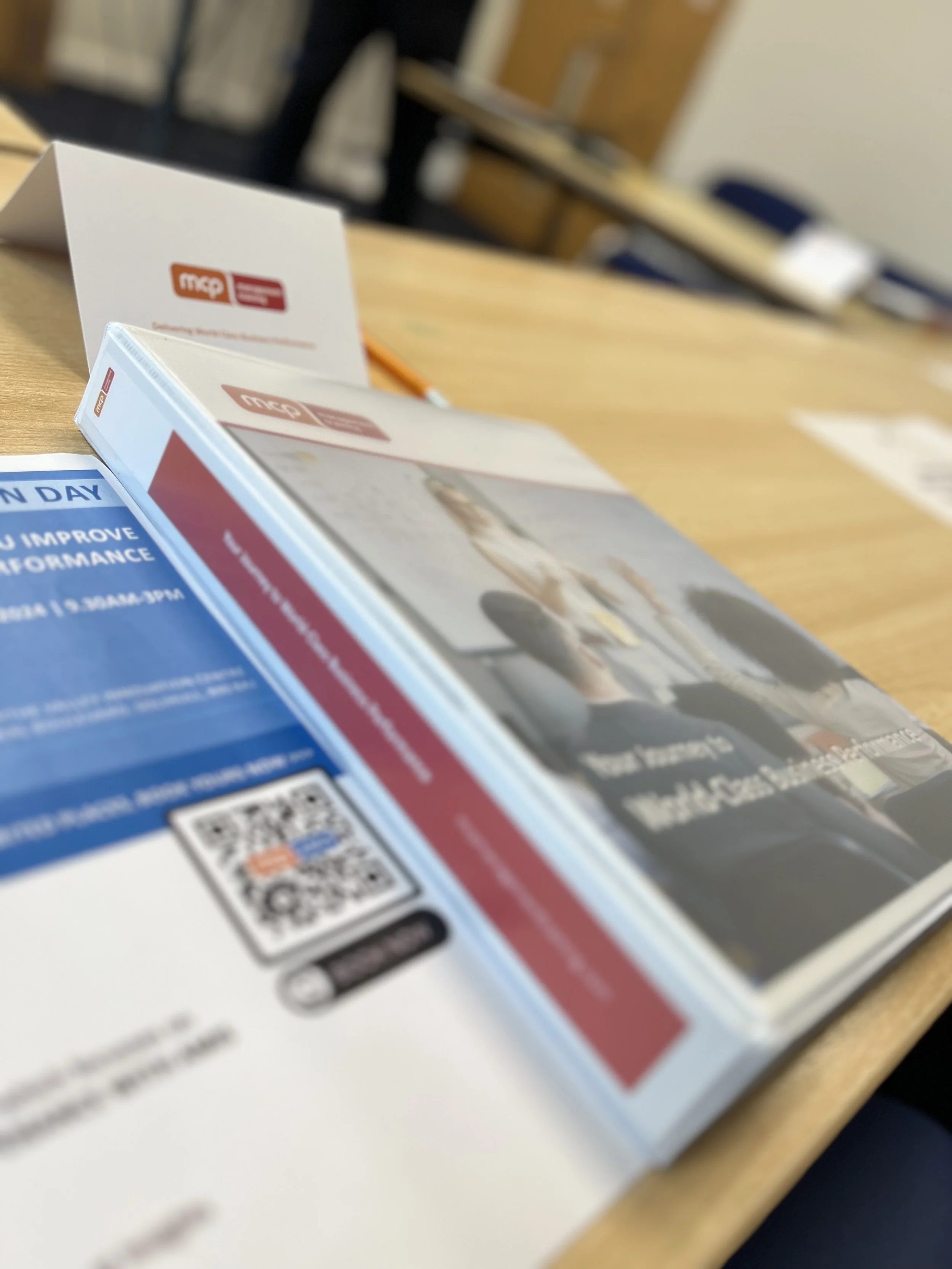 Close-up of a desk with pamphlets and flyers, one of which has text about performance improvement and a QR code, office setting in the background.