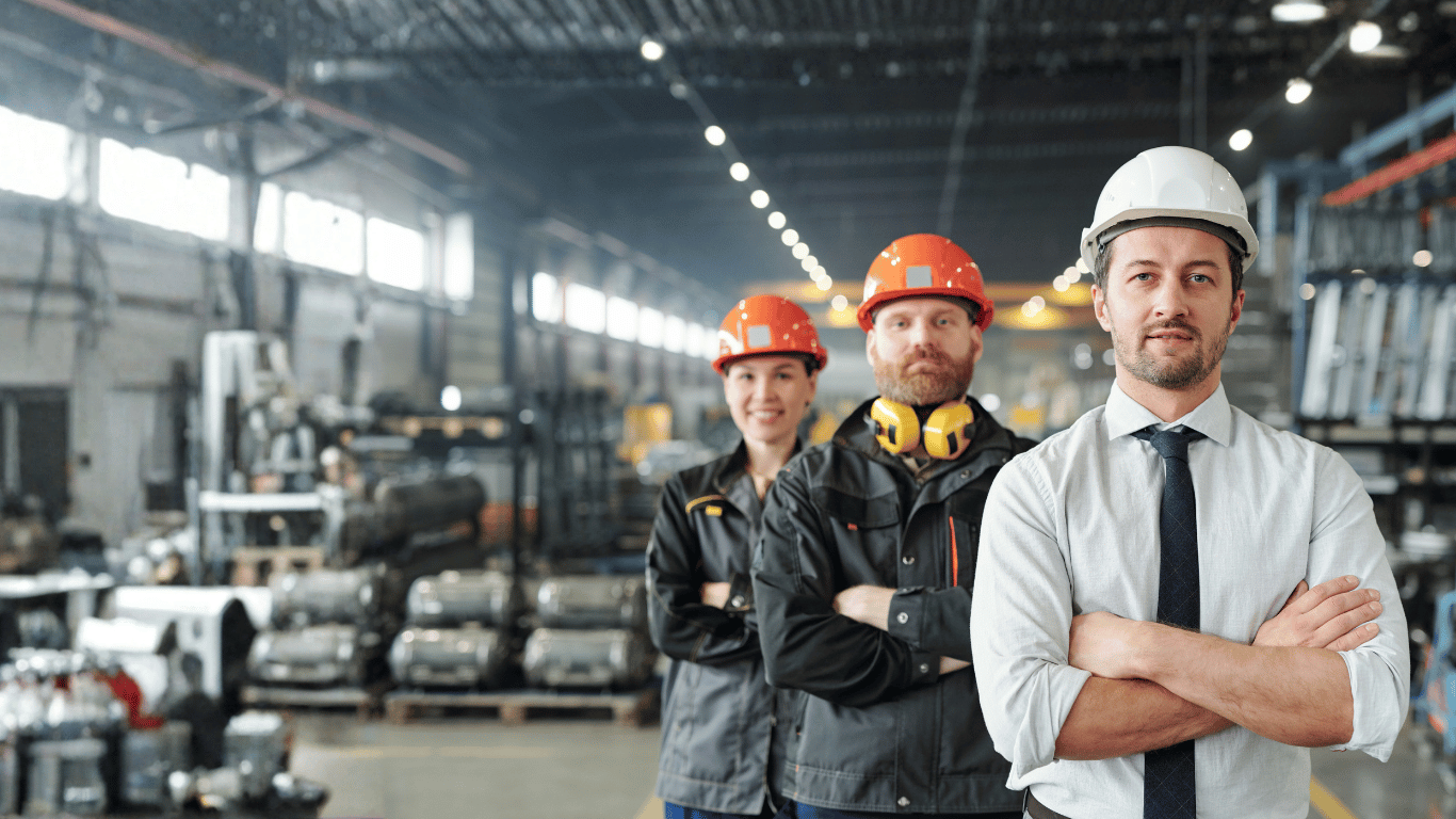 Three factory workers, two men and one woman, standing with arms crossed inside a large industrial warehouse. They are wearing safety helmets, with the woman and one man in orange helmets and the other man in a white helmet. The warehouse has shelves and equipment in the background.