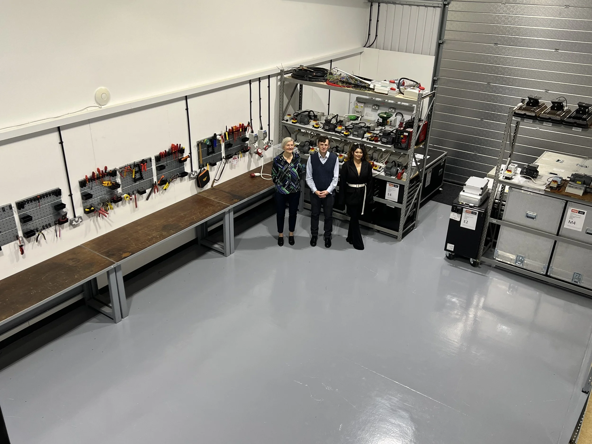 Three people standing in a workshop with tools on the wall and electronic equipment on shelves around them.