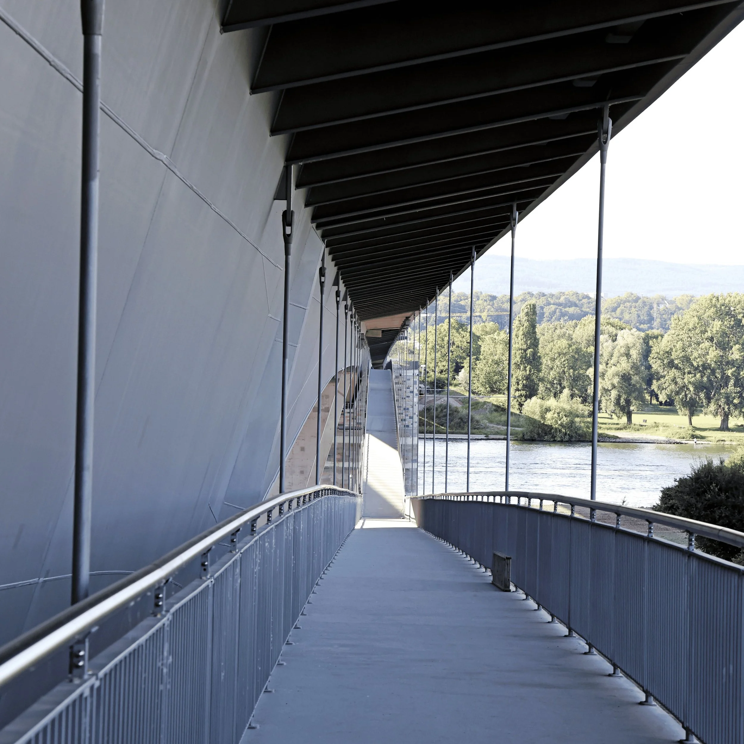 Blick entlang einer modernen Brücke mit überdachtem Gehweg, Geländern und einer Flusslandschaft im Hintergrund.