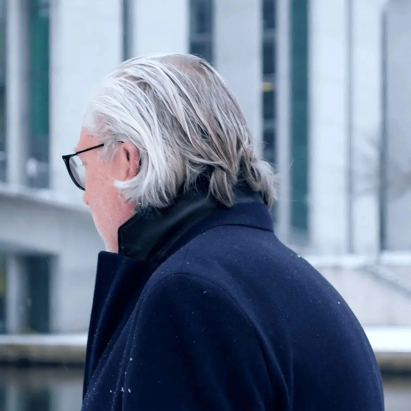 Ein älterer Mann mit langen, silbergrauen Haaren und Brille, gekleidet in einen dunklen Mantel, steht draußen vor einem modernen Gebäude.